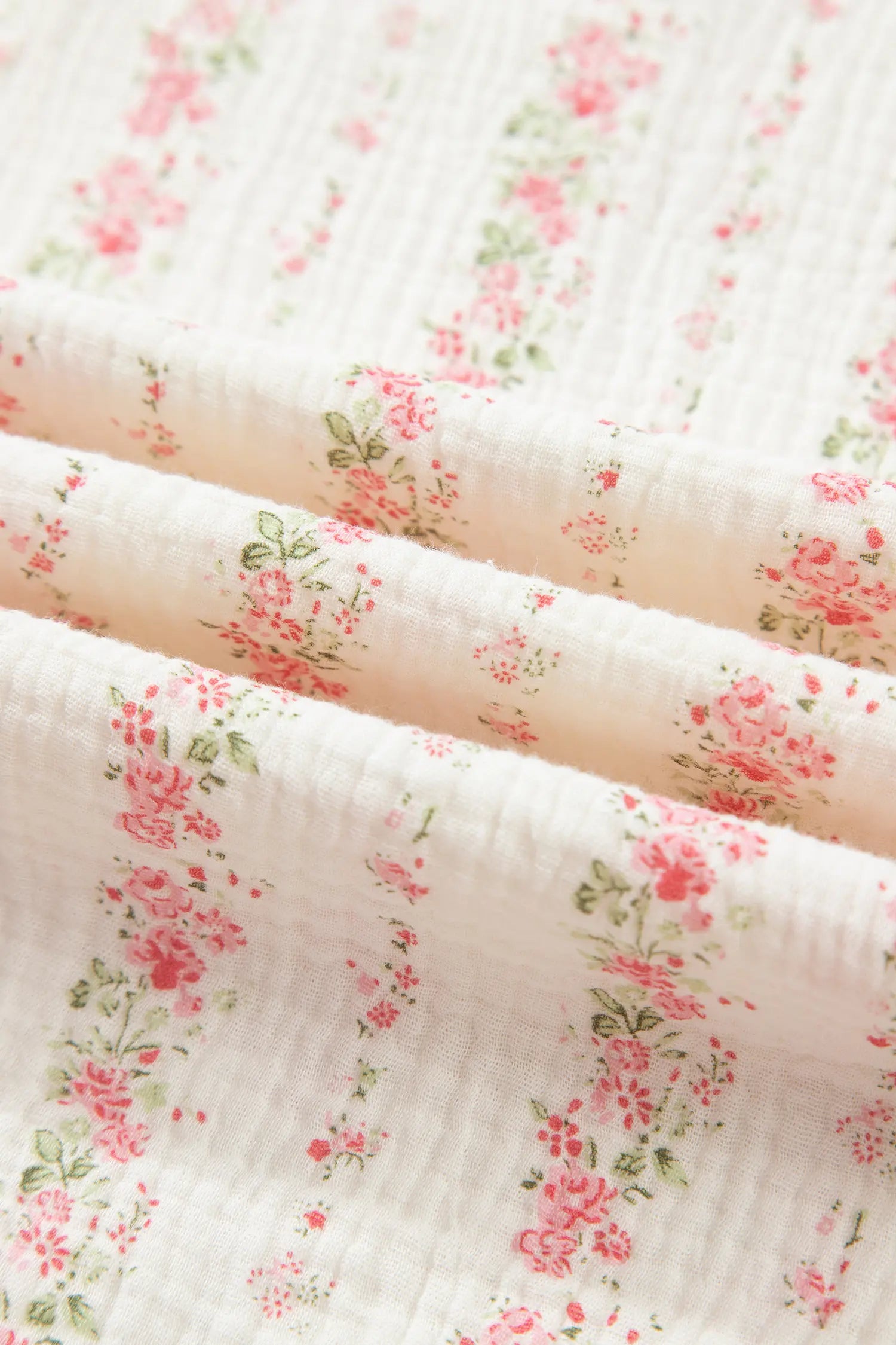 A close-up of a textured muslin fabric with a delicate pink and green floral print on a warm off-white background.