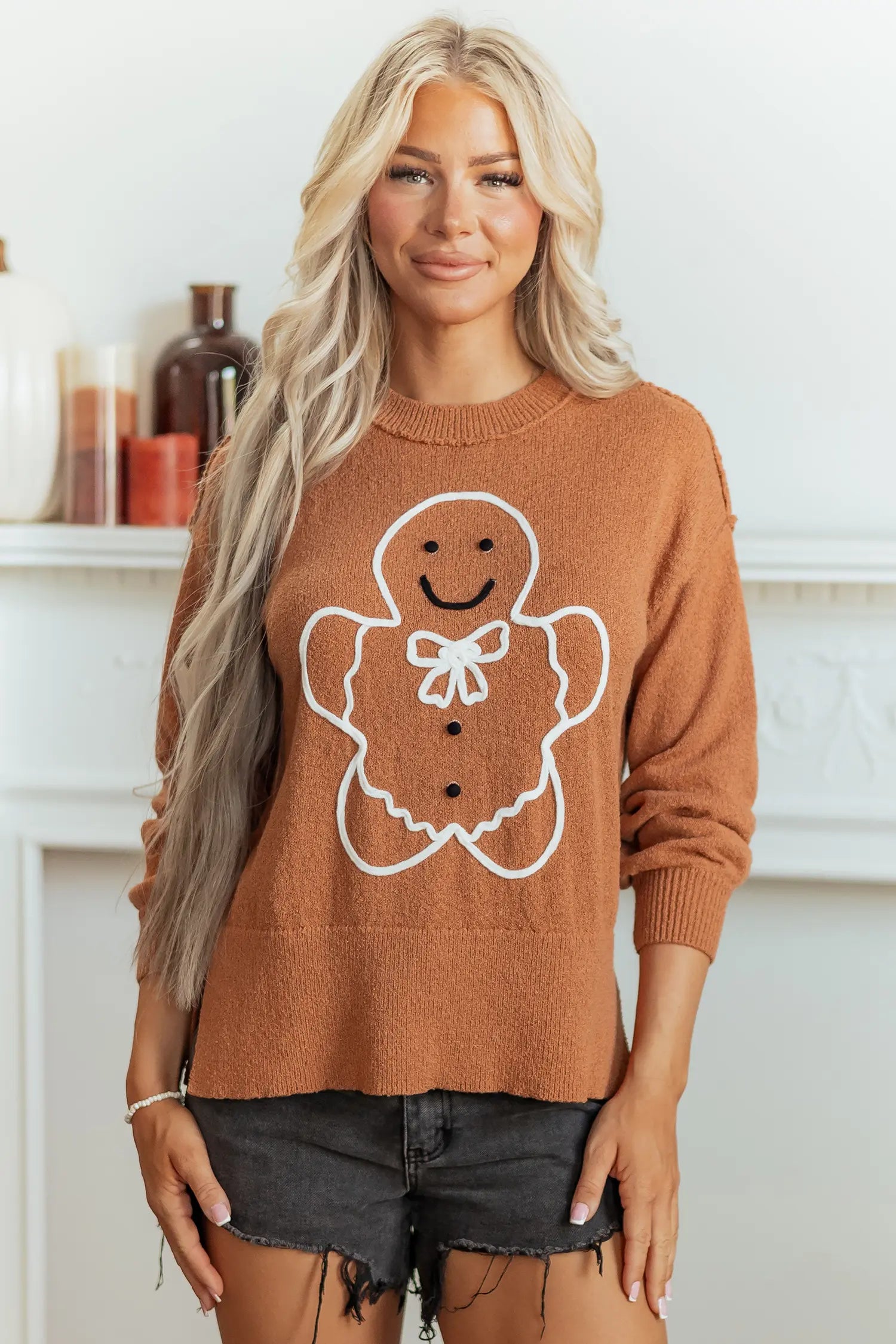 A woman wearing a rust-orange crewneck sweater with a white gingerbread man graphic on the front, paired with dark denim shorts, standing in front of a white fireplace mantel.
