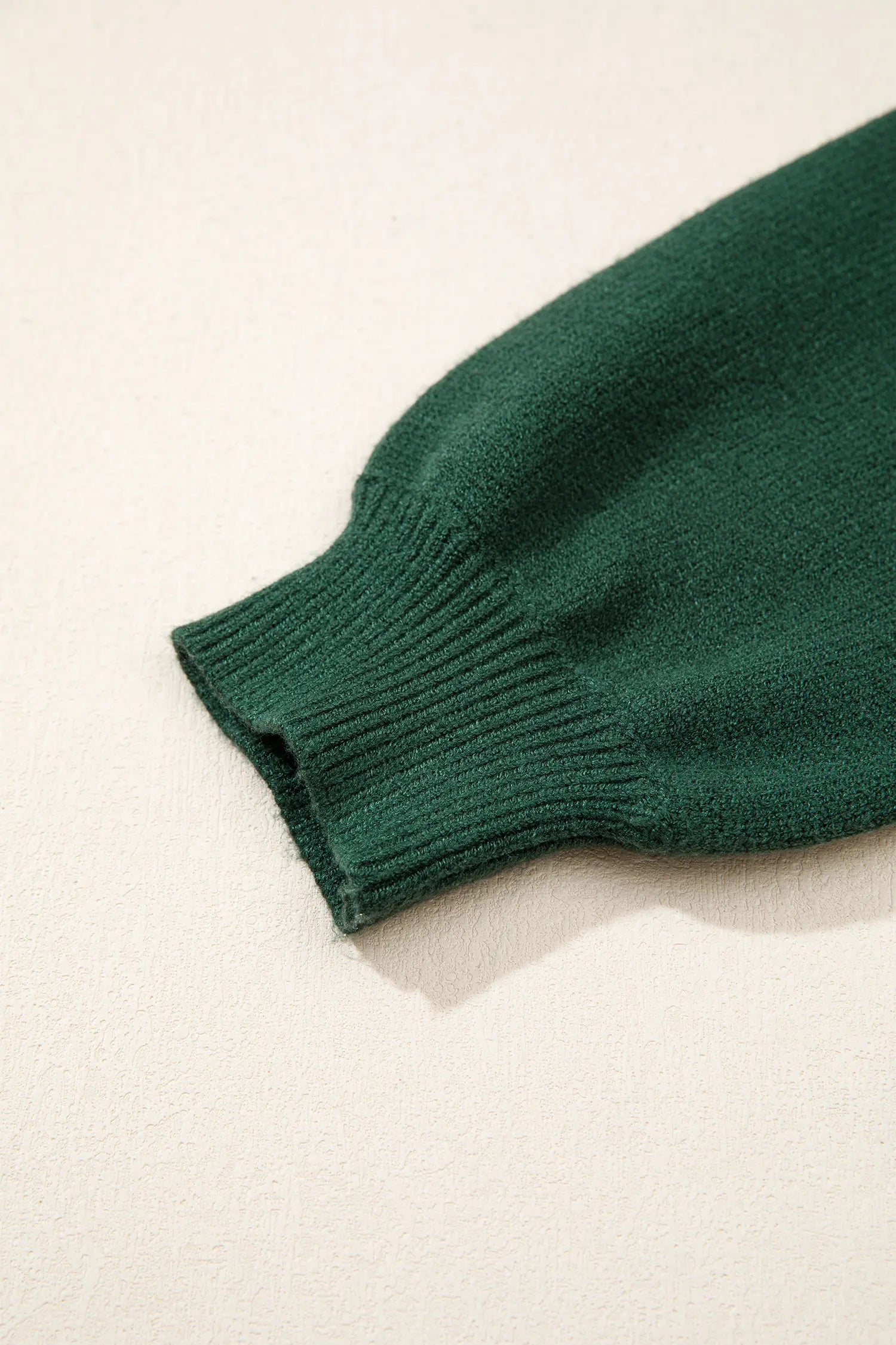 Close-up of a dark forest green ribbed knit sleeve or cuff on a light cream textured background.