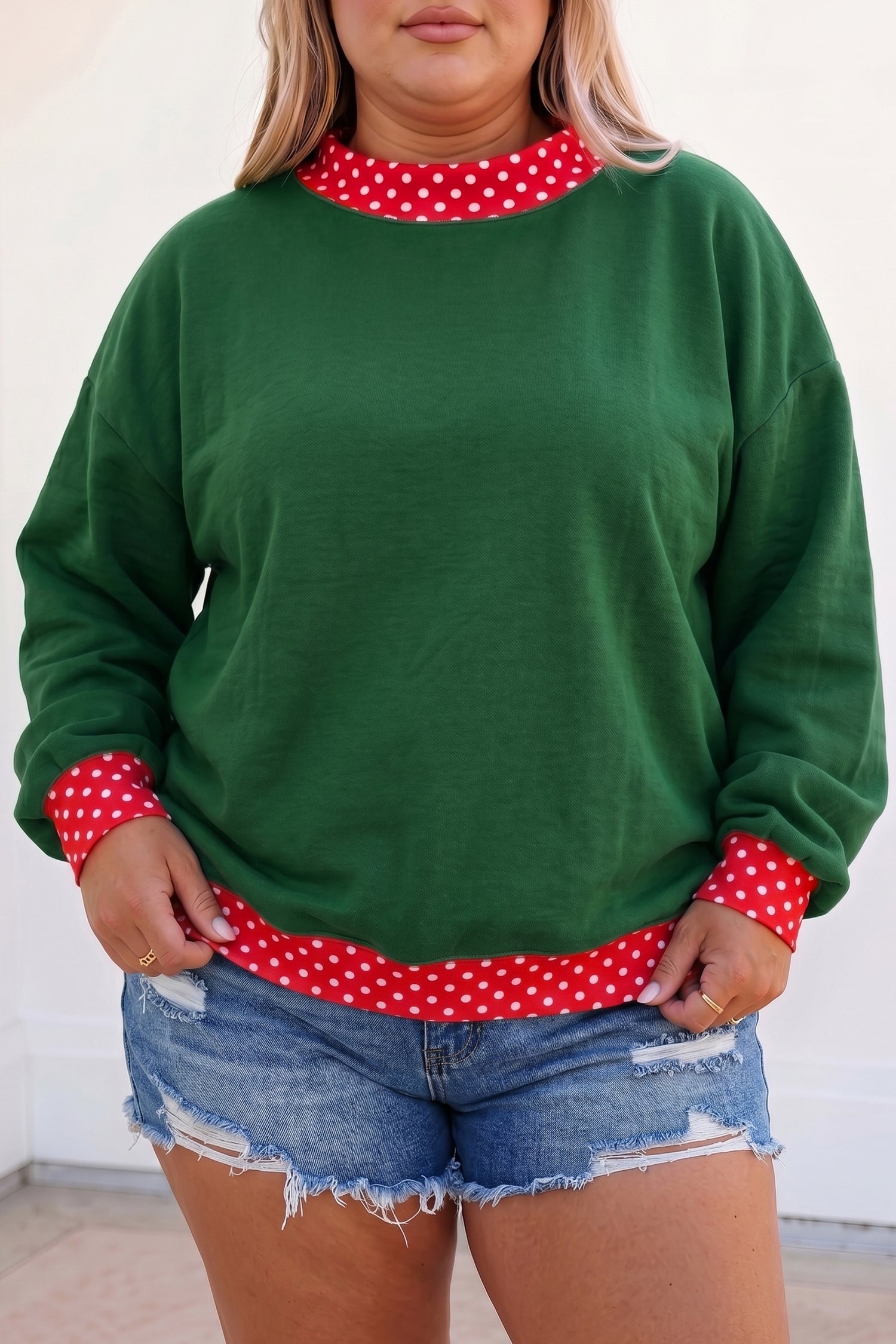A woman wearing a green crewneck sweatshirt with red and white polka dot trim at the collar, cuffs, and hem, paired with denim shorts.