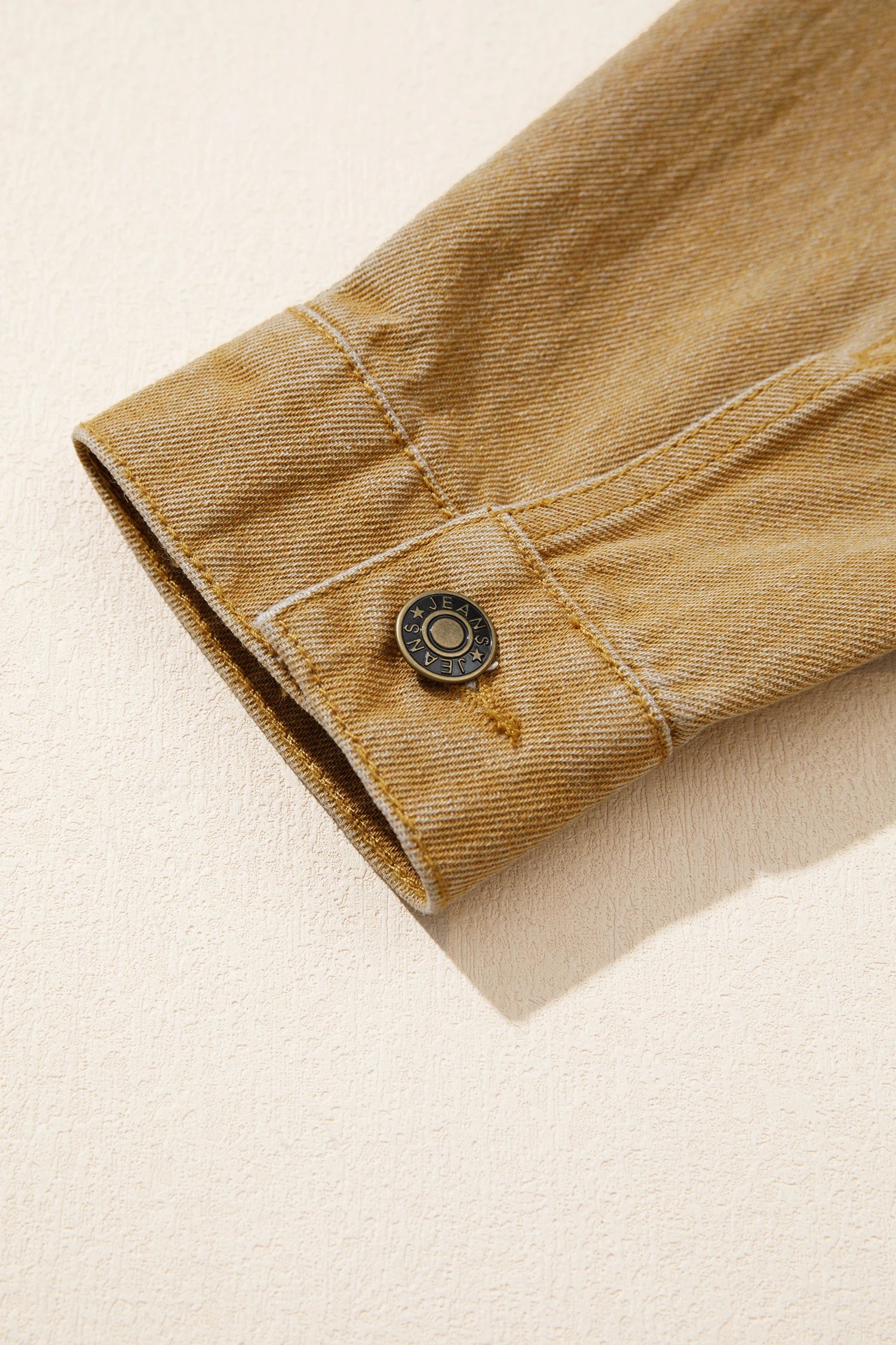 Close-up of a tan denim jacket sleeve cuff with a single round metal button, set against a textured cream background.