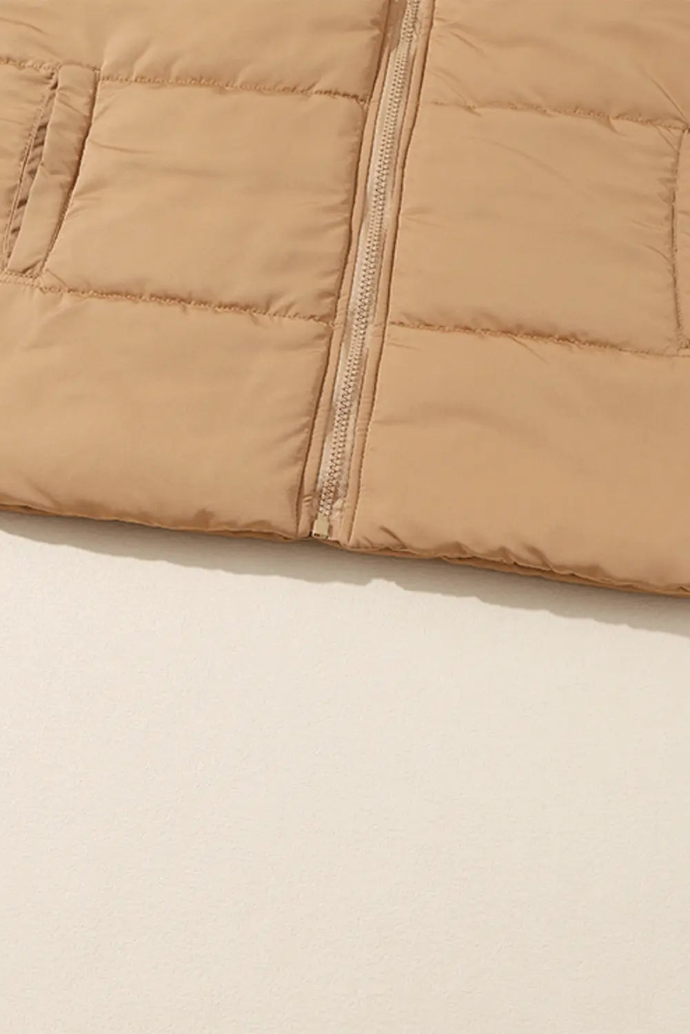 Close-up of beige quilted puffer jacket with front zipper and padded panels against a light background.