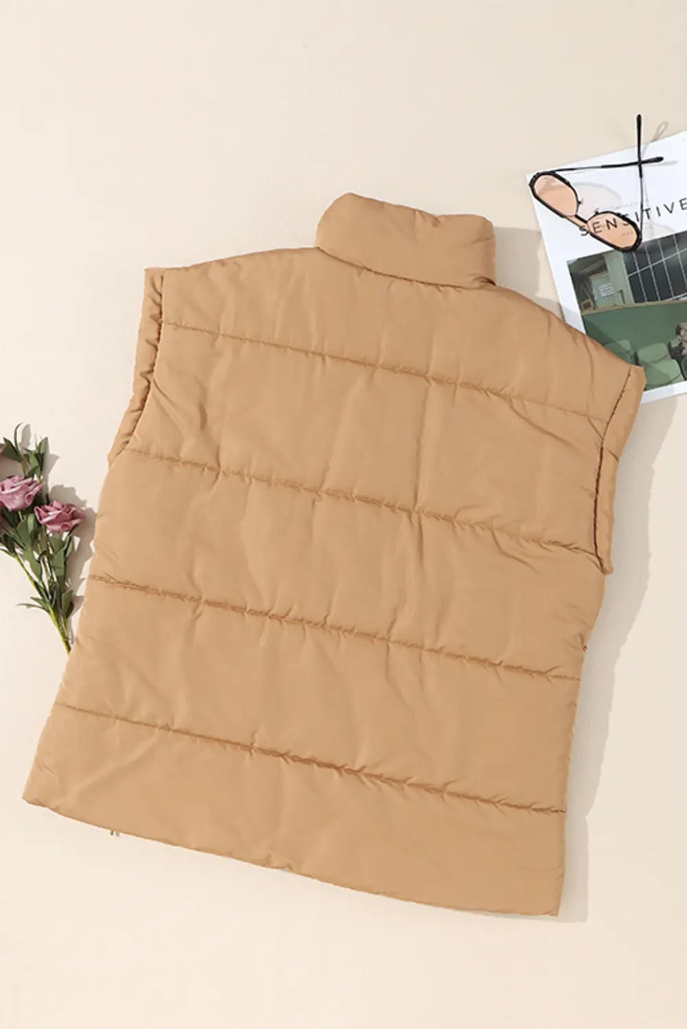 Camel tan sleeveless quilted puffer vest laid flat with flower sprig and magazine on beige background.