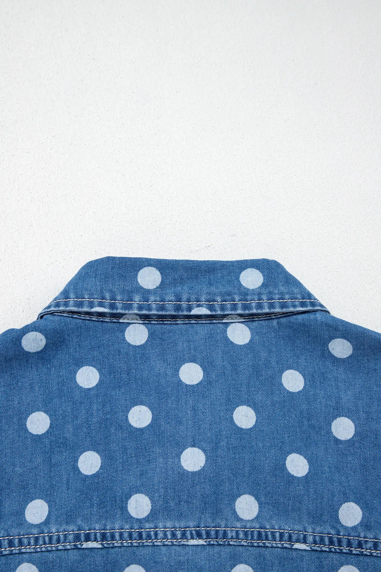 Close-up of blue denim shirt with white polka dots, showing collar and upper back against a white background.