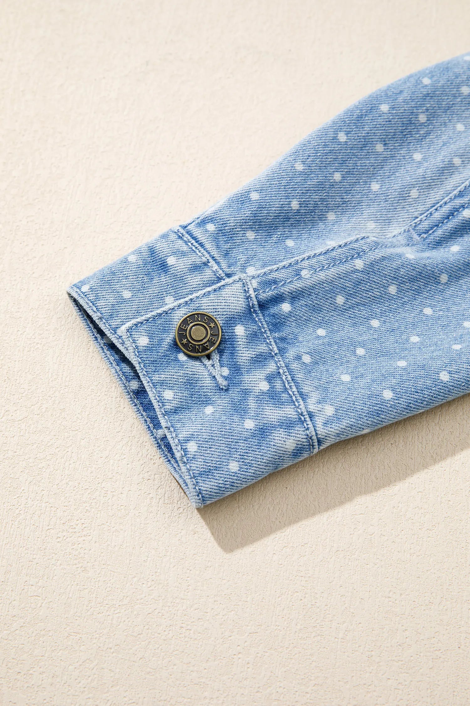 Close-up of light blue denim sleeve with white polka dots and dark metal button cuff.