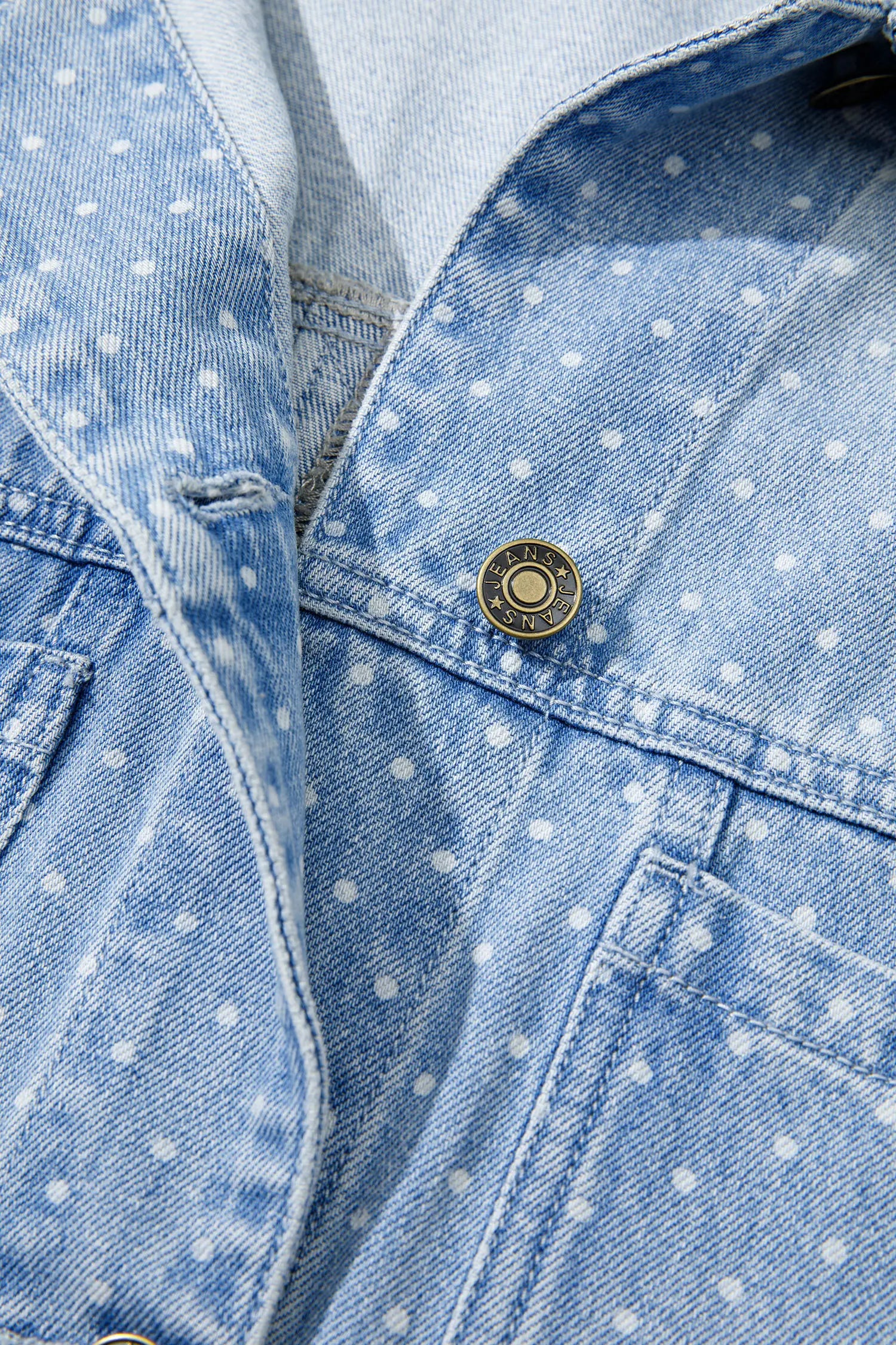 Close-up of a light blue denim jacket with a white polka dot pattern, featuring a brass button closure.