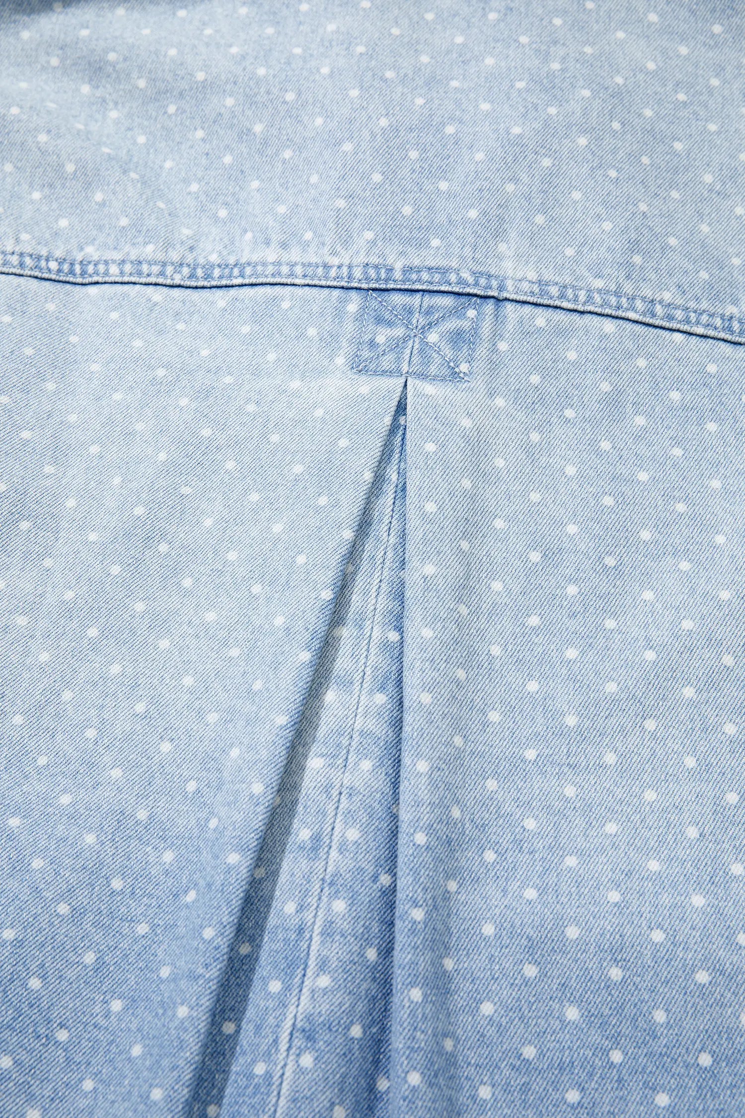 Close-up of light blue denim with white polka dots, stitched seam, and small square patch.