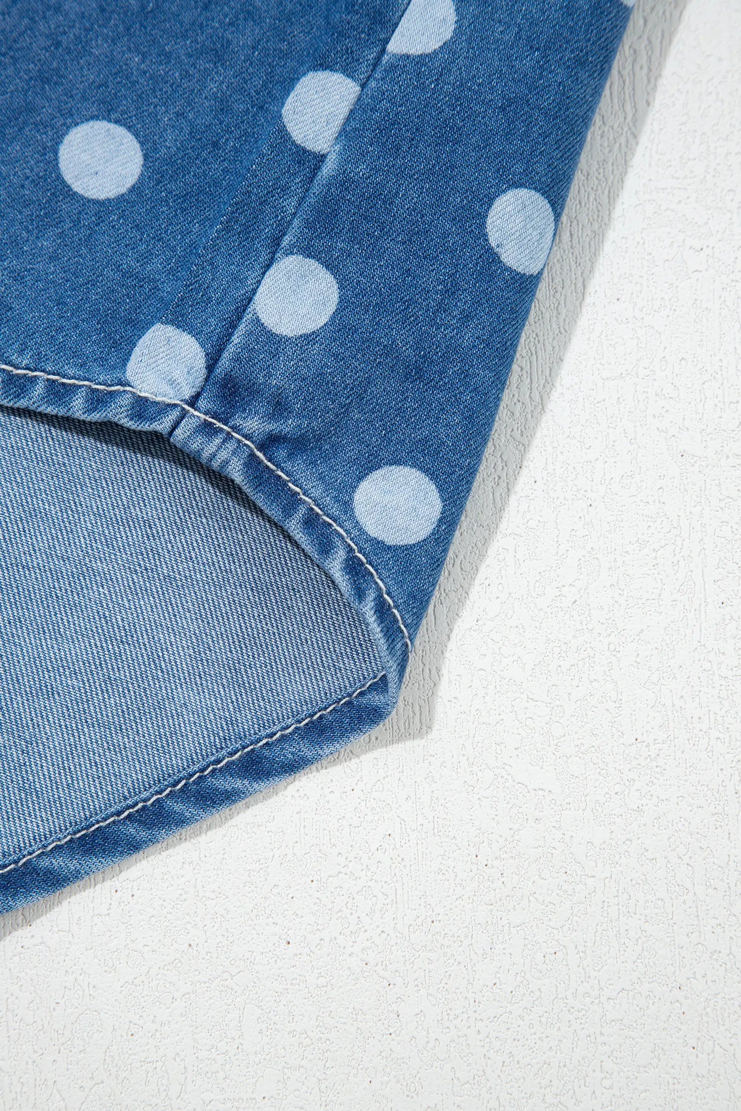 Close-up of blue denim with white polka dots, showing textured fabric and folded hem on an off-white background.