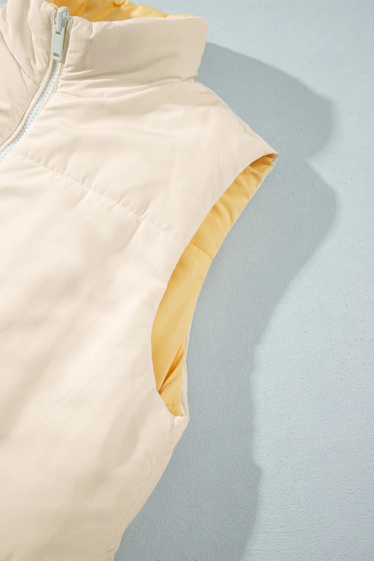 Close-up of light cream puffer vest with yellow lining, high collar, and light blue zipper.
