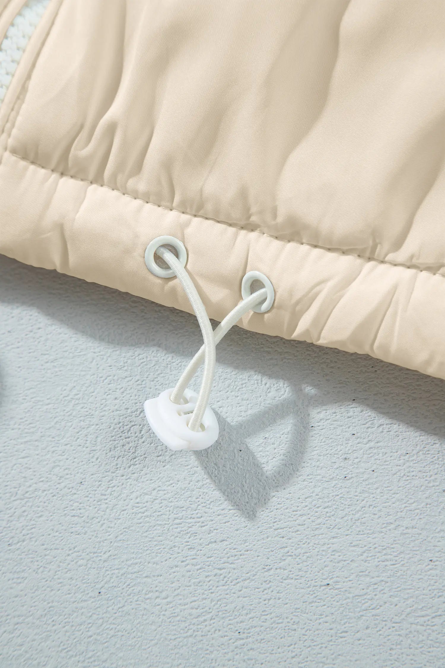 Close-up of quilted beige puffer jacket with white drawstrings and blue eyelets against gray-blue wall.