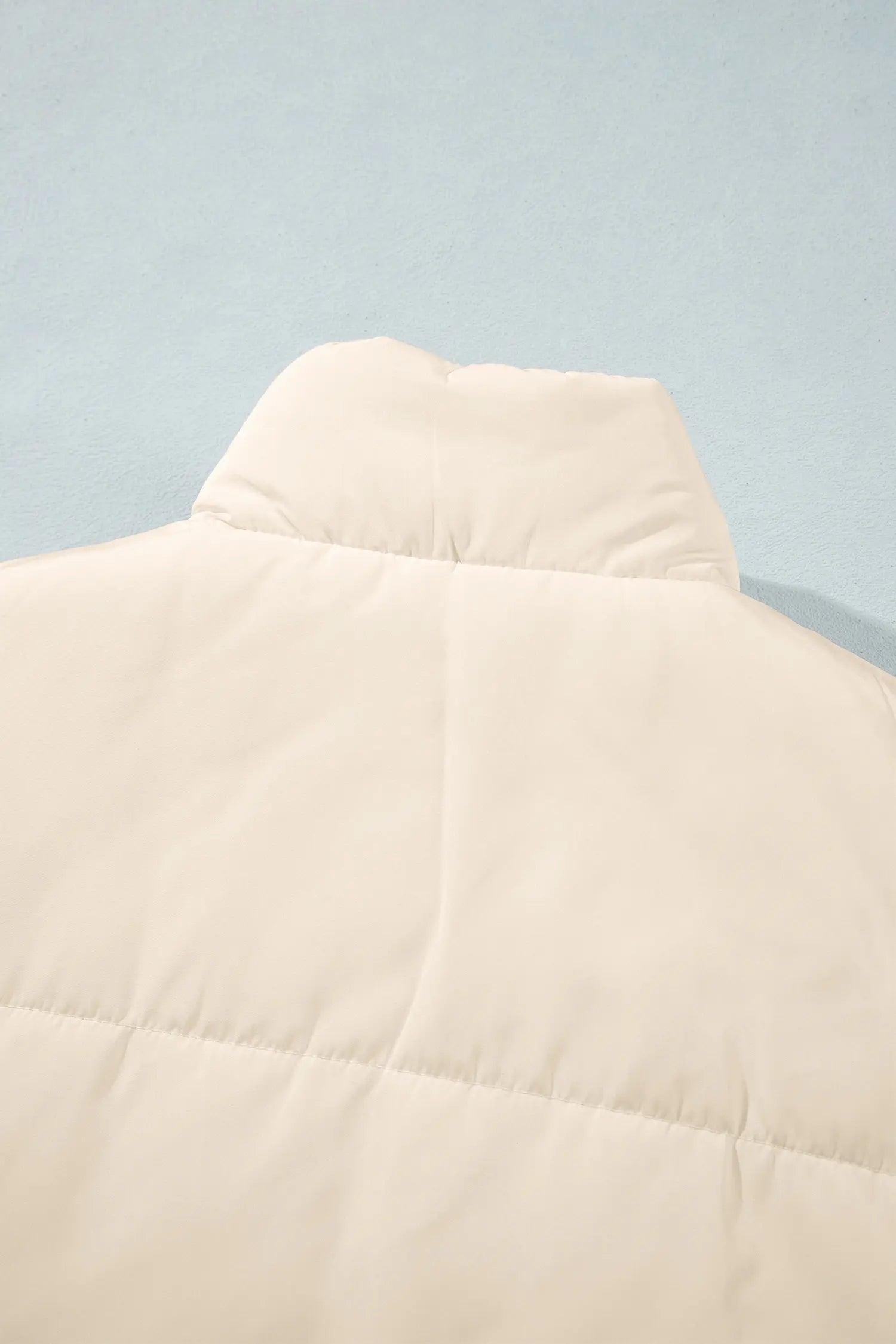 Close-up of cream-colored puffer jacket with padded collar and horizontal quilting on light blue background.