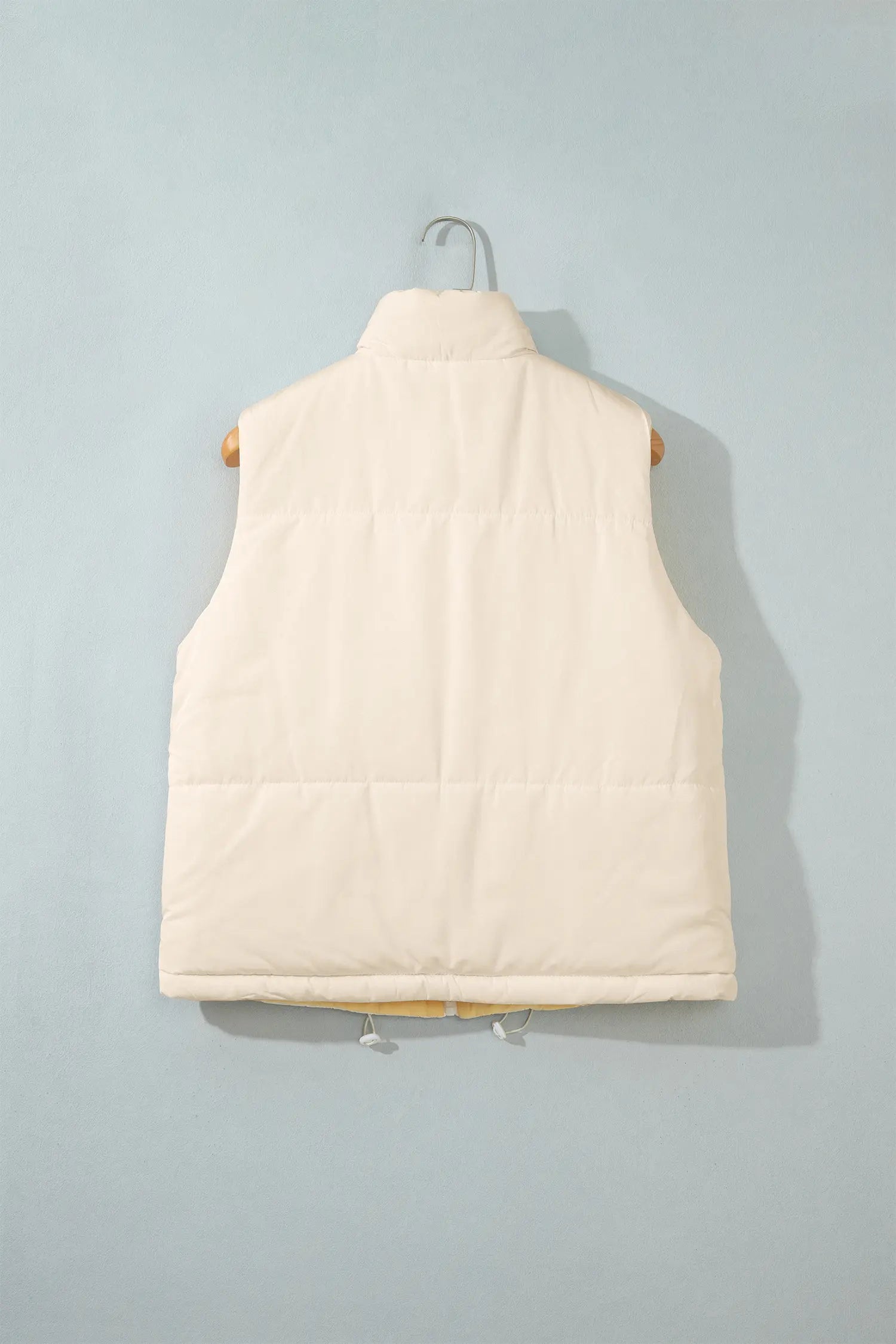 Cream-colored sleeveless puffer vest hanging on a wooden hanger against a light blue-gray background.