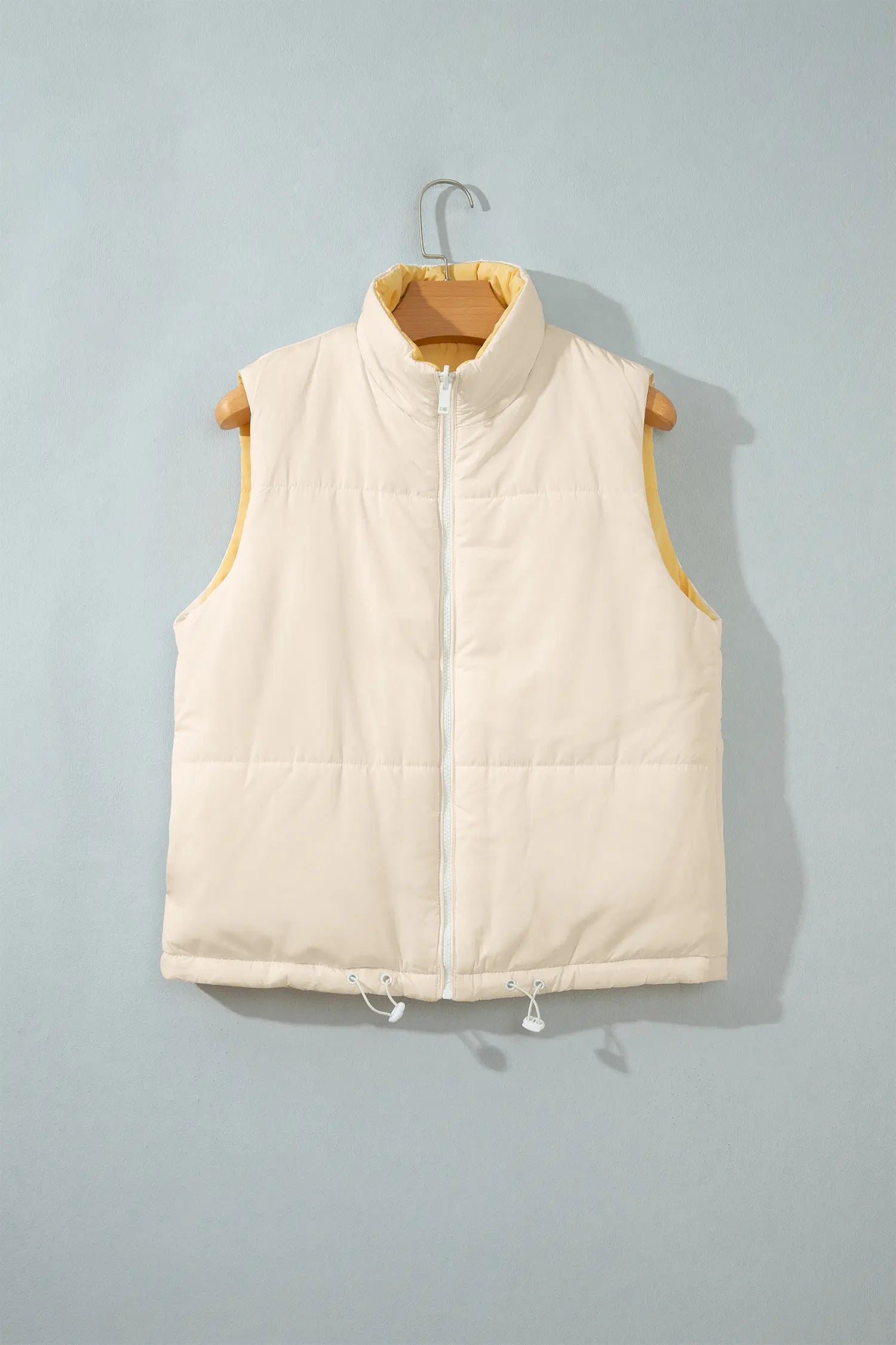 Cream sleeveless puffer vest with stand-up collar and white zipper on a wooden hanger.