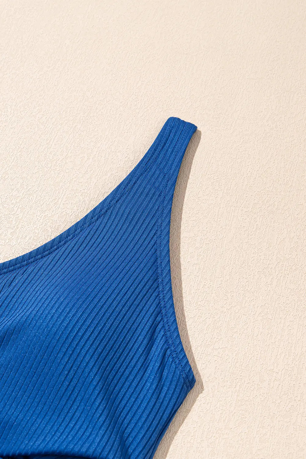 A close-up of a bright blue ribbed knit top or bralette strap against a warm beige textured background.
