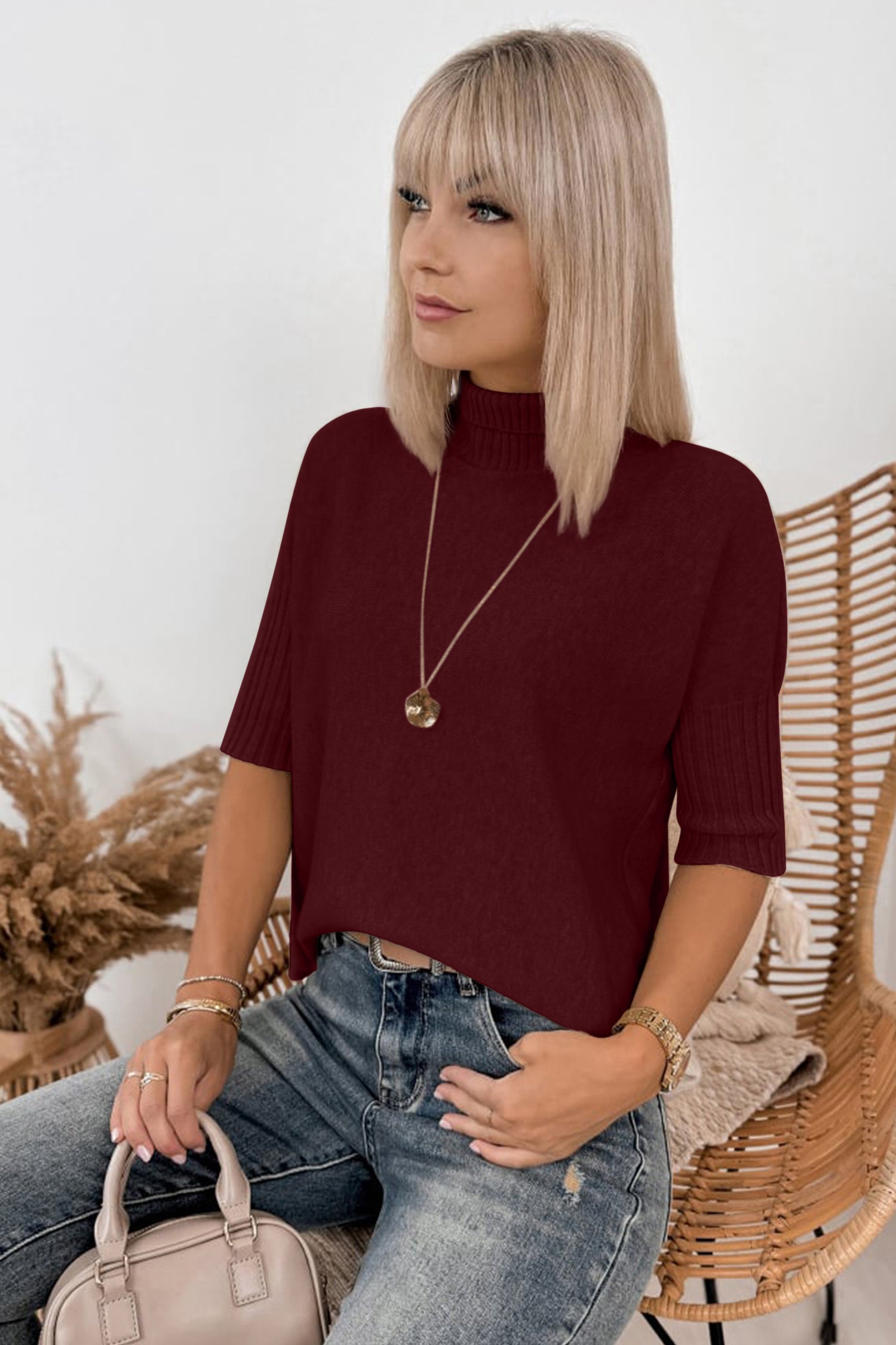 Blonde woman in a burgundy short-sleeve ribbed mock neck top and blue jeans on a wicker chair.