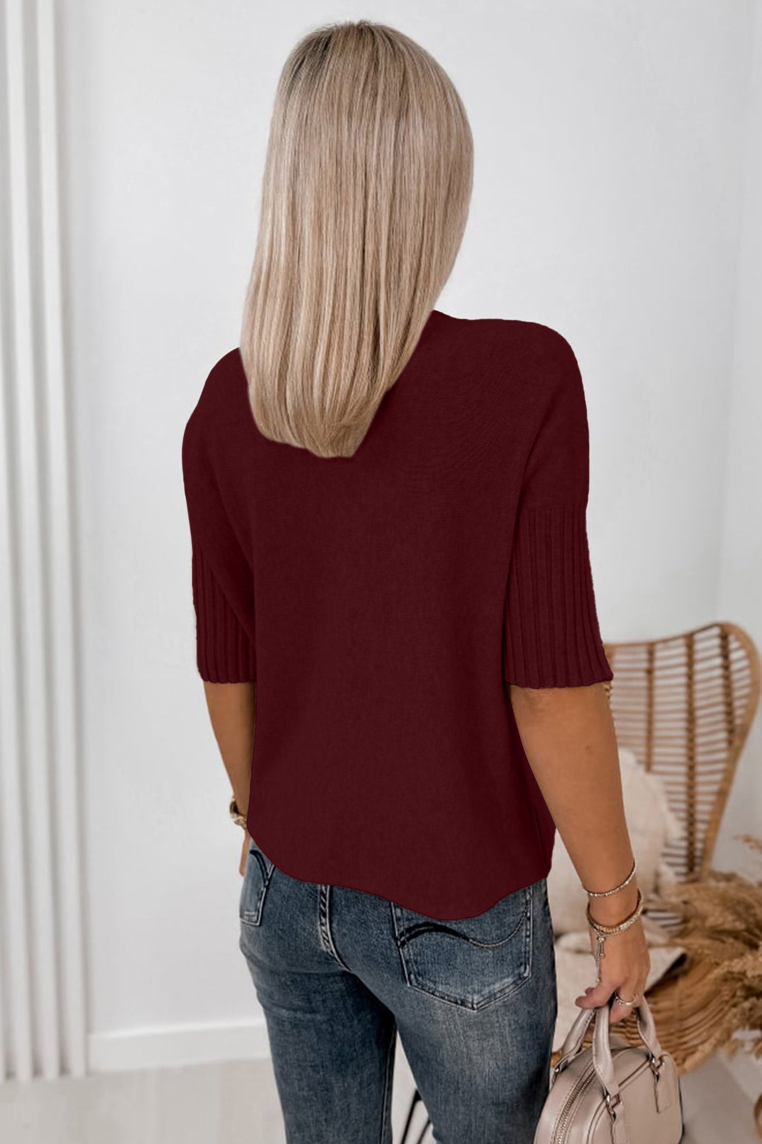 Woman from behind in a burgundy short-sleeve knit top, blue jeans, holding a light beige bag.