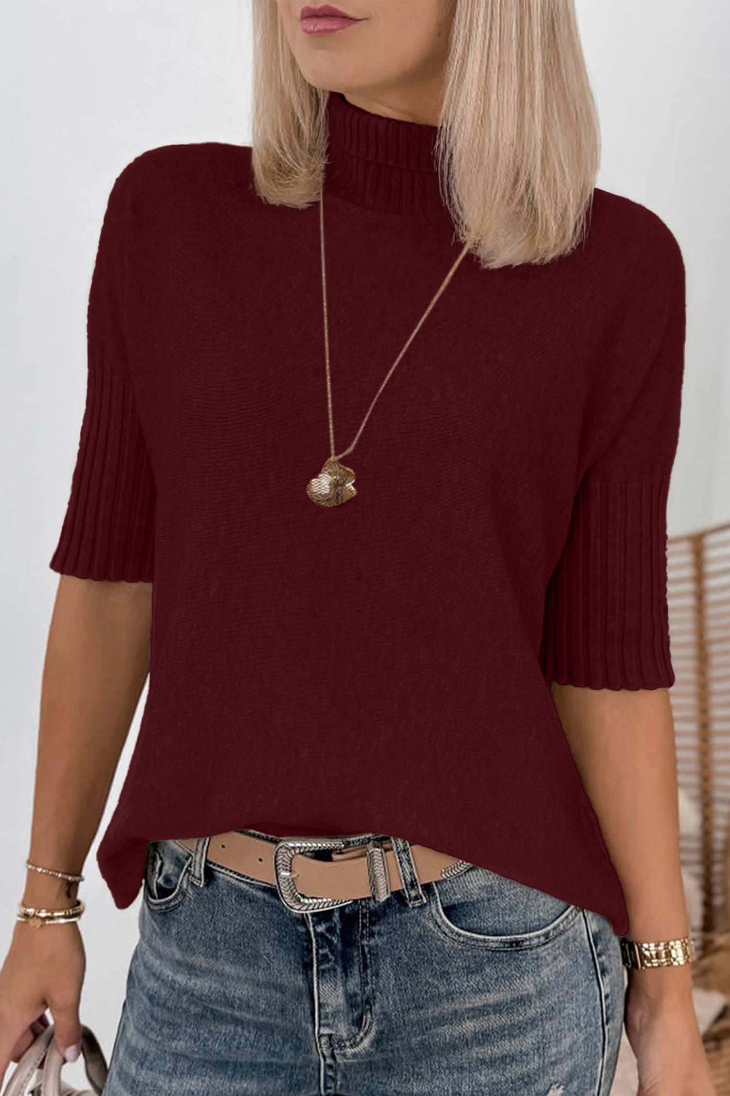 Woman in burgundy short-sleeve mock neck knit sweater, blue jeans, and gold pendant necklace.