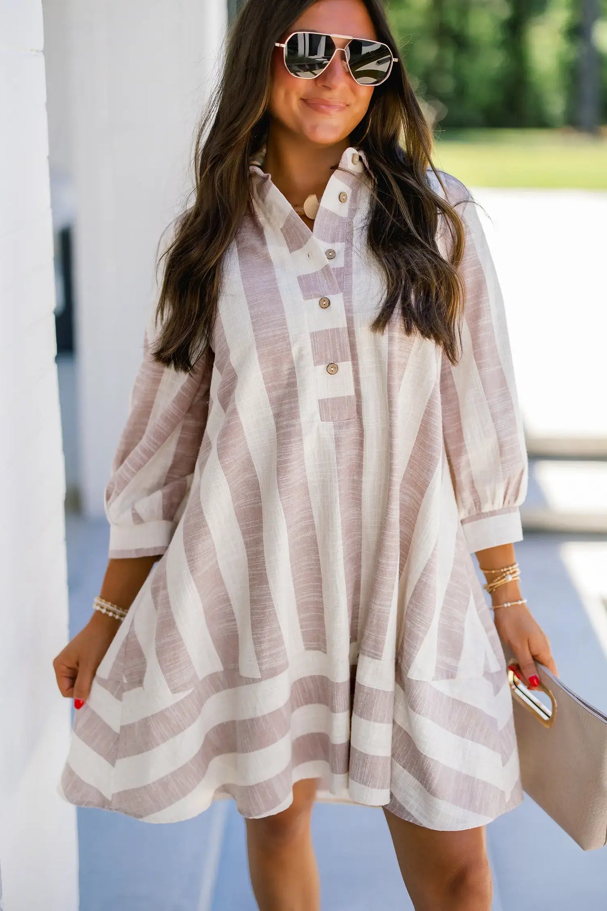 Woman wearing a striped dress with sunglasses and holding a clutch outdoors.