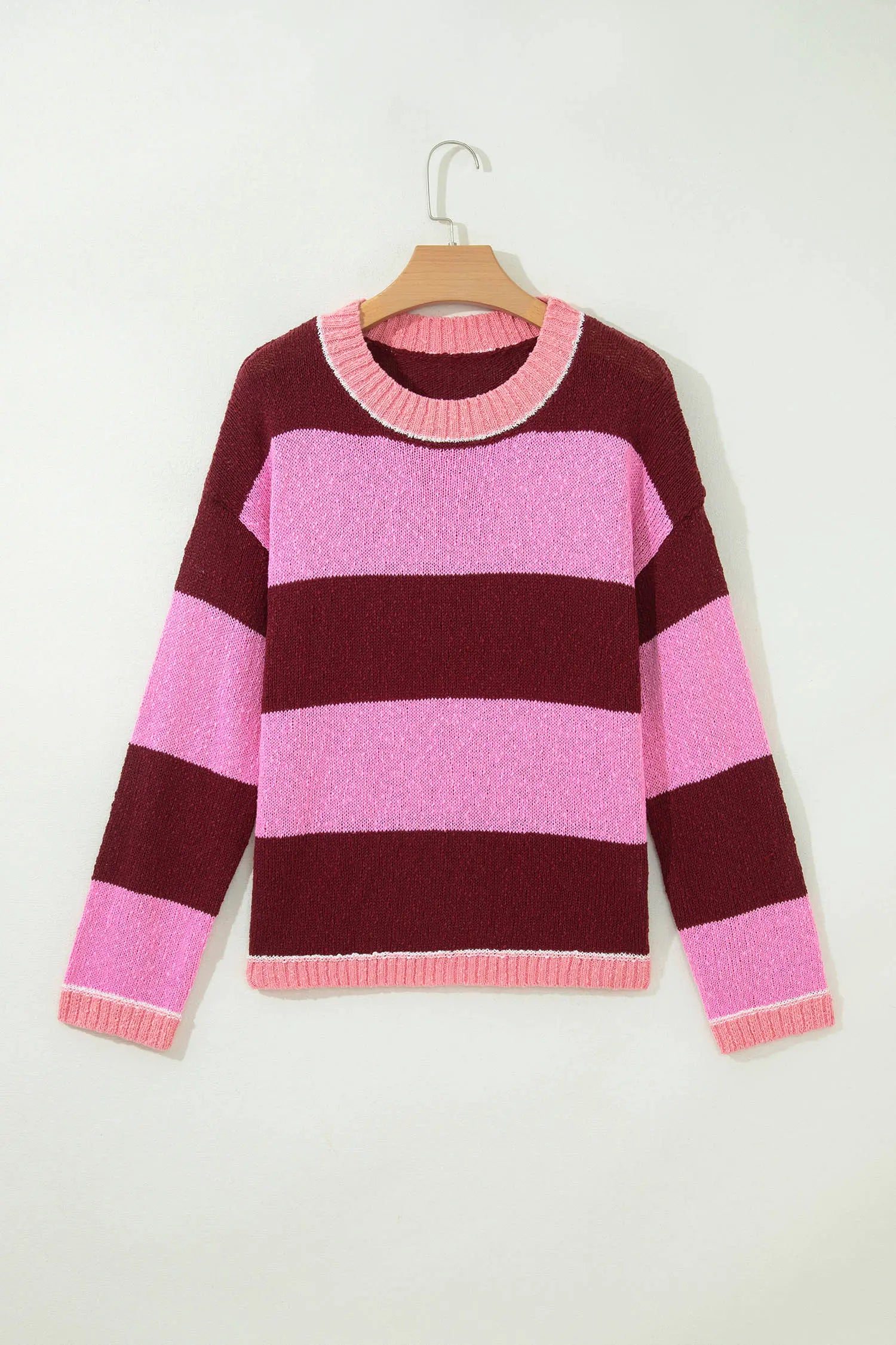 A pink and burgundy striped knit sweater hanging on a wooden hanger against a plain off-white wall.