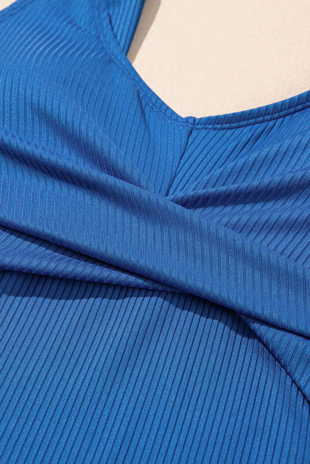 Close-up of a blue ribbed knit top with a twisted crossover neckline, shown on a mannequin against a light beige background.