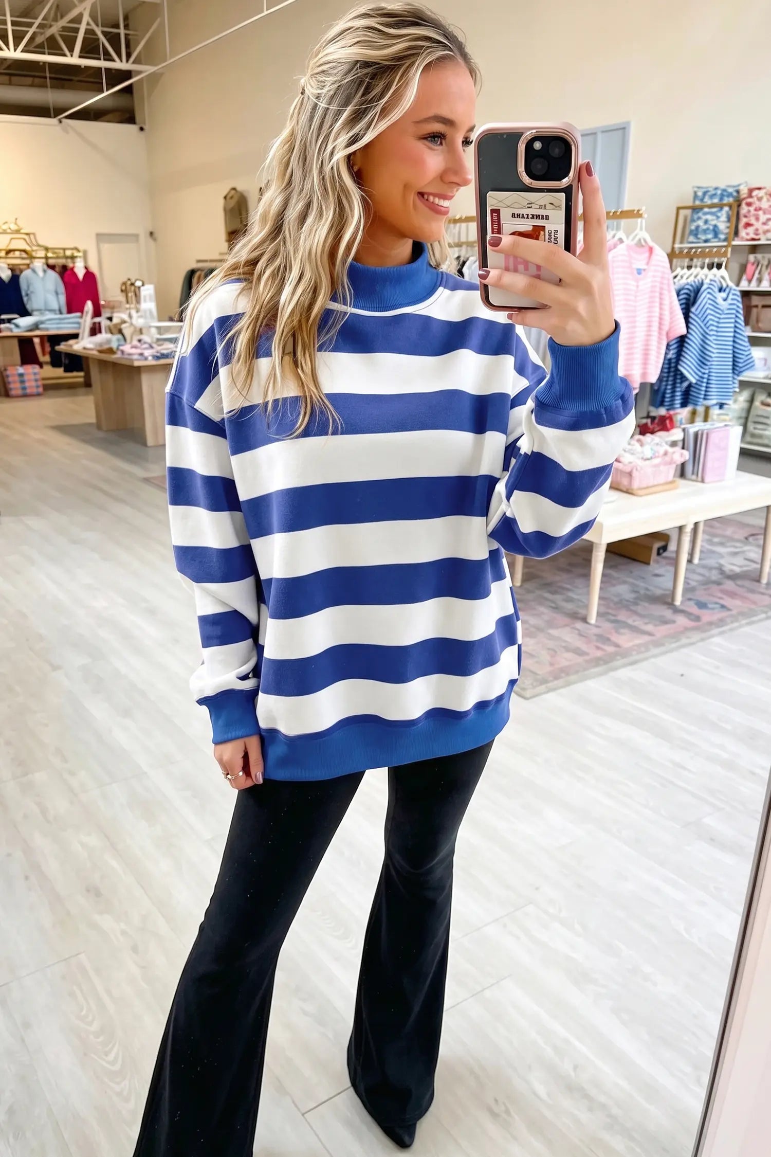 A woman takes a mirror selfie in a clothing store, wearing a blue and white horizontally striped mock neck sweatshirt and black flare pants.