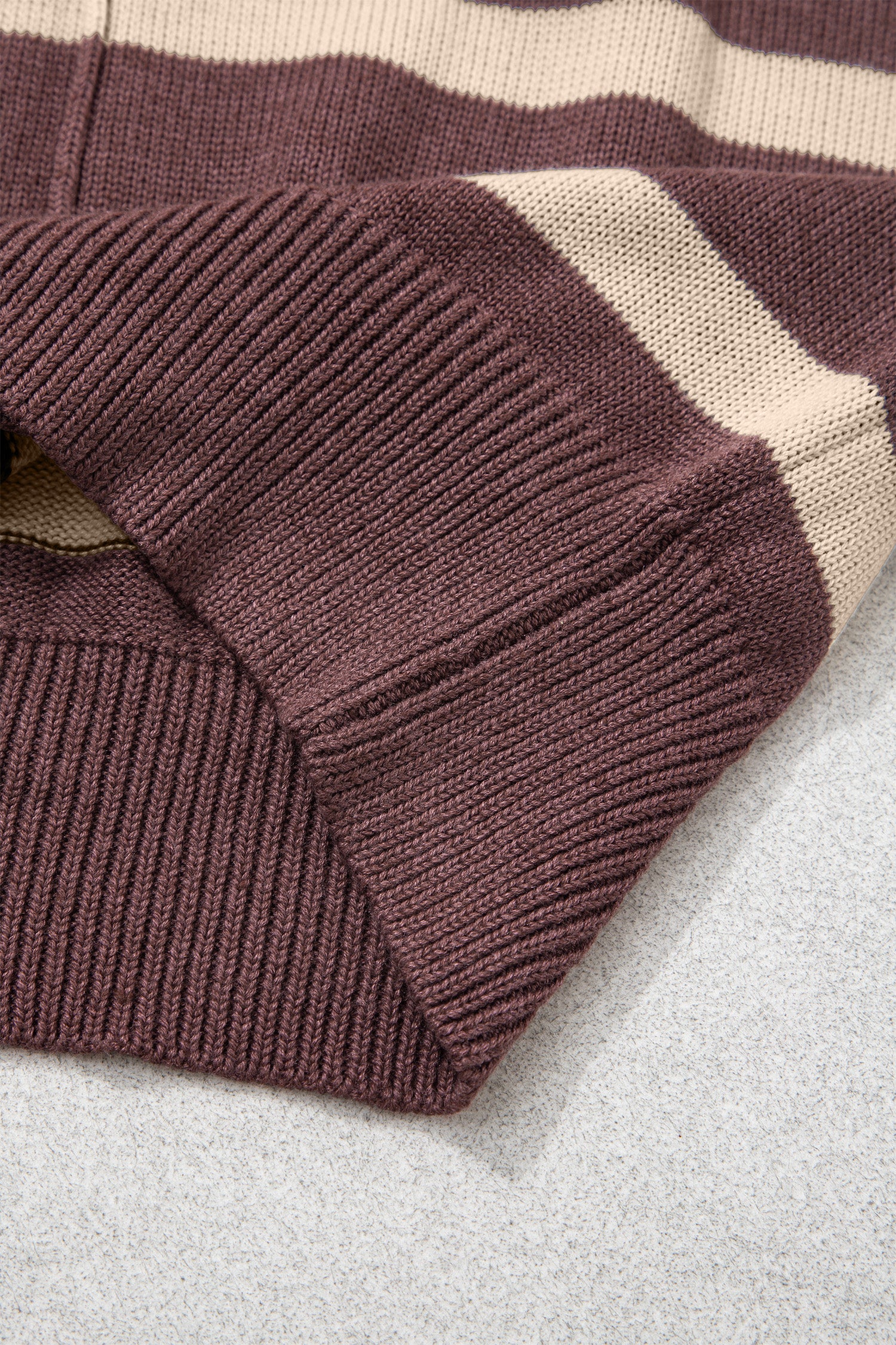 Close-up of a ribbed knit sweater corner showing a deep burgundy ribbed cuff and body with horizontal cream stripes, laid on a light gray textured surface.