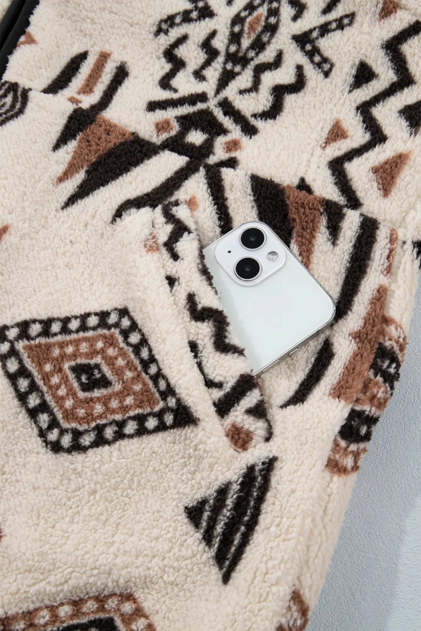 Close-up of cream sherpa fleece garment with black-brown geometric pattern and a white smartphone in the pocket.