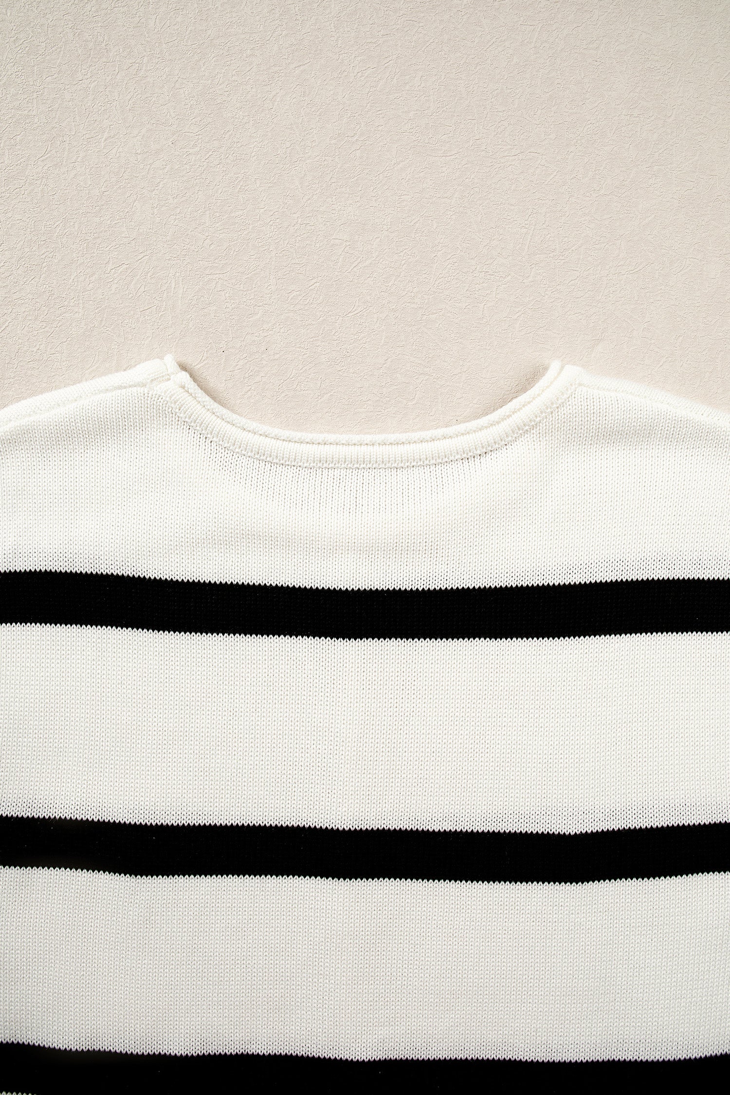 Close-up of a white knit sweater with horizontal black stripes, showing the crew neck and ribbed texture against a light beige background.