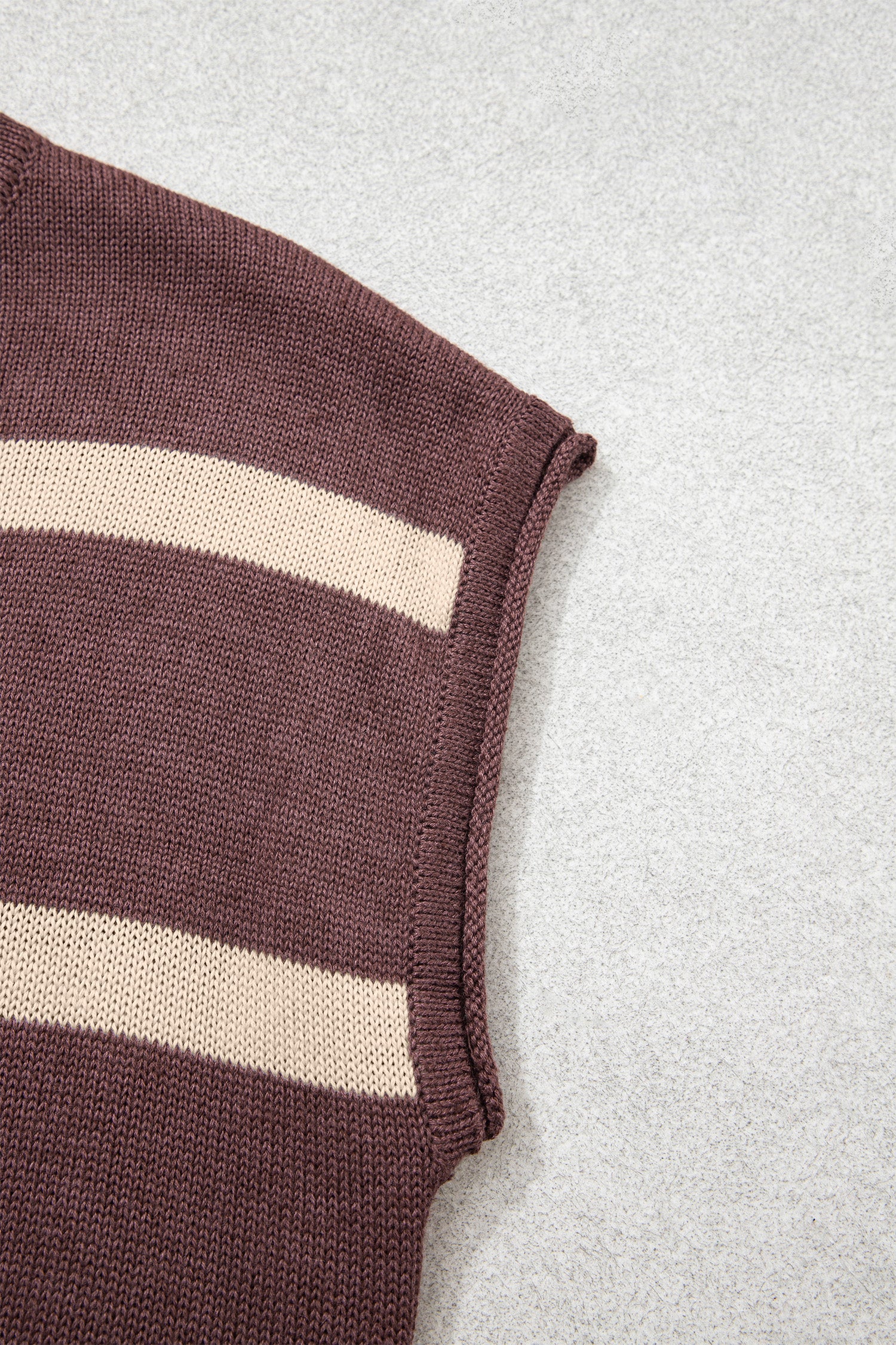 Close-up of a ribbed knit sleeve or cuff in dark brown with two horizontal cream stripes, laid on a light gray textured background.