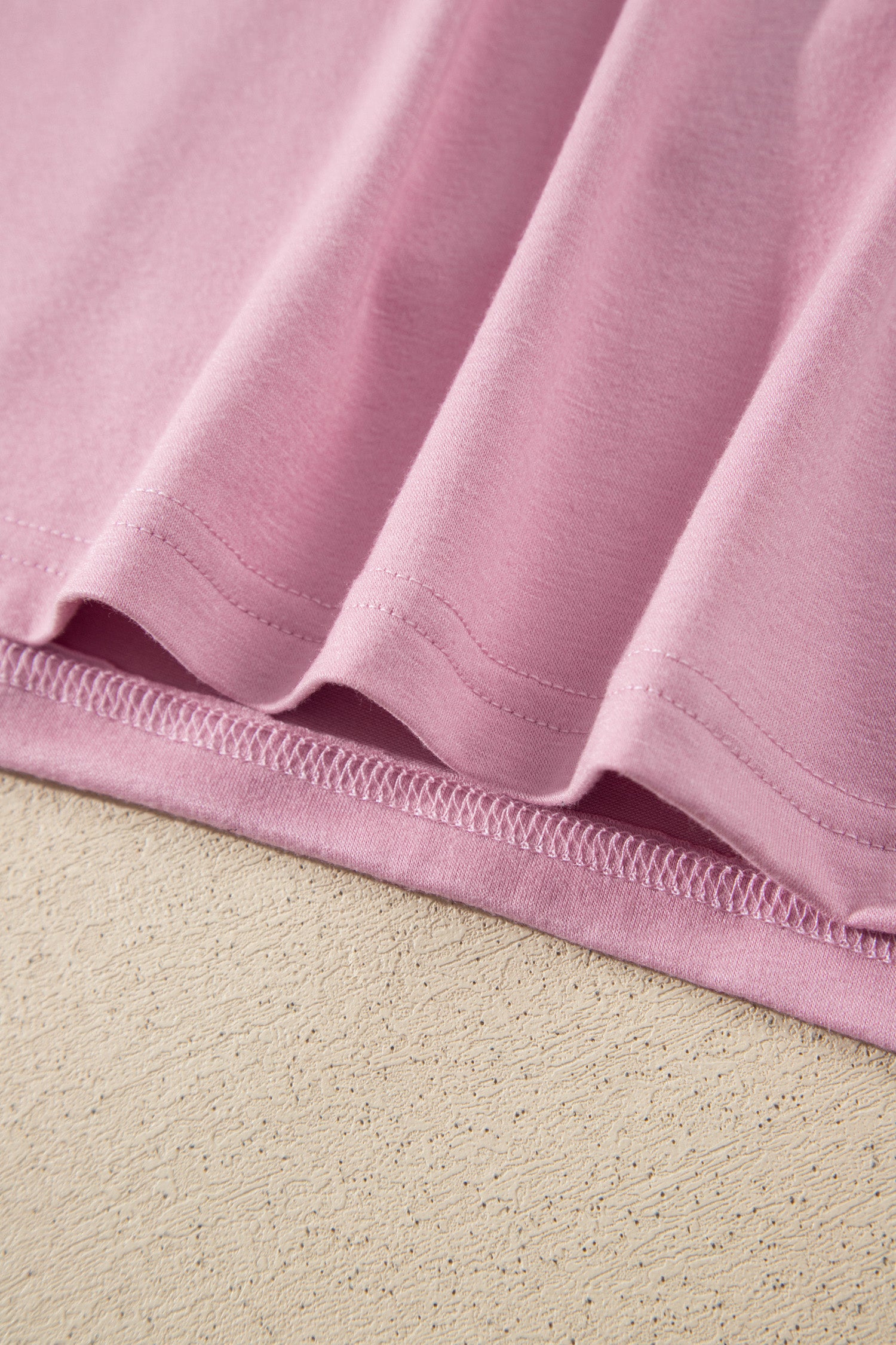 Close-up of a dusty pink t-shirt hem with neat double stitching, laid flat on a textured beige surface.