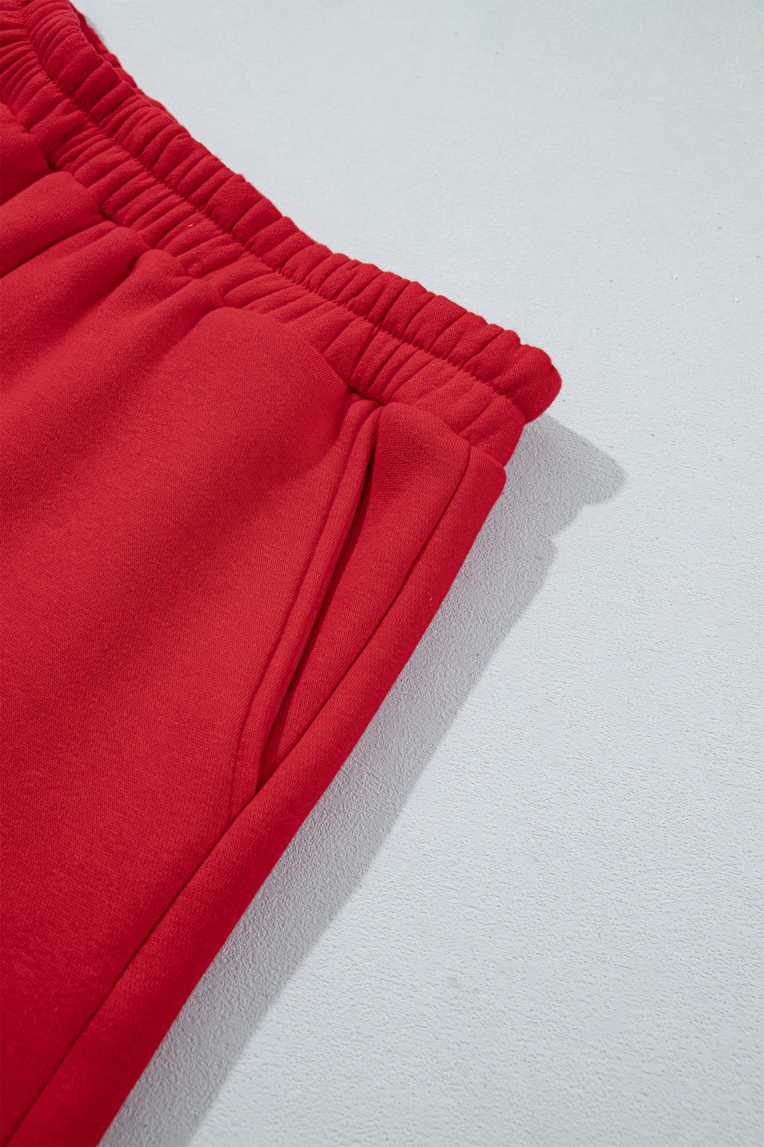 A close-up detail shot of bright red sweatpants, showing the elastic waistband and side pocket on a light gray textured background.