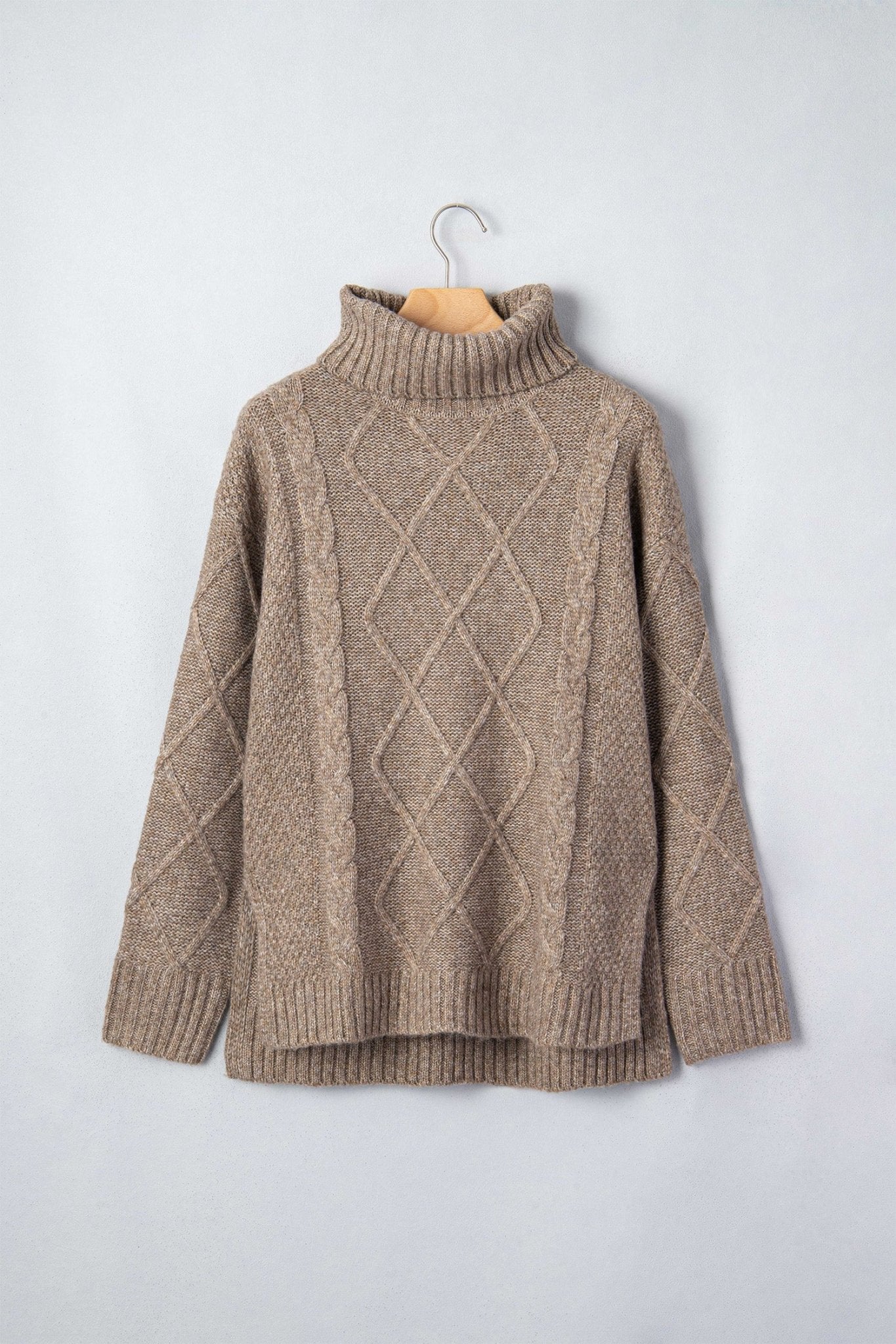 A beige cable-knit turtleneck sweater hanging on a wooden hanger against a light gray wall.