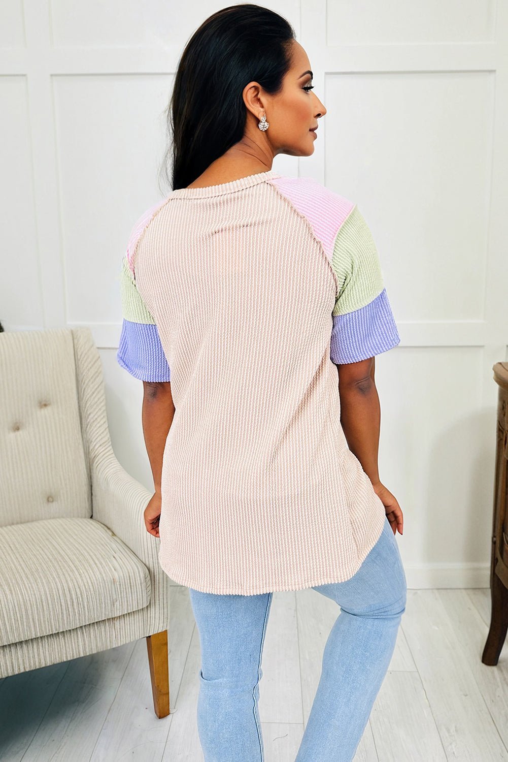 Woman wearing a pastel color-blocked top and light blue jeans in a room with a white wall and gray chair.