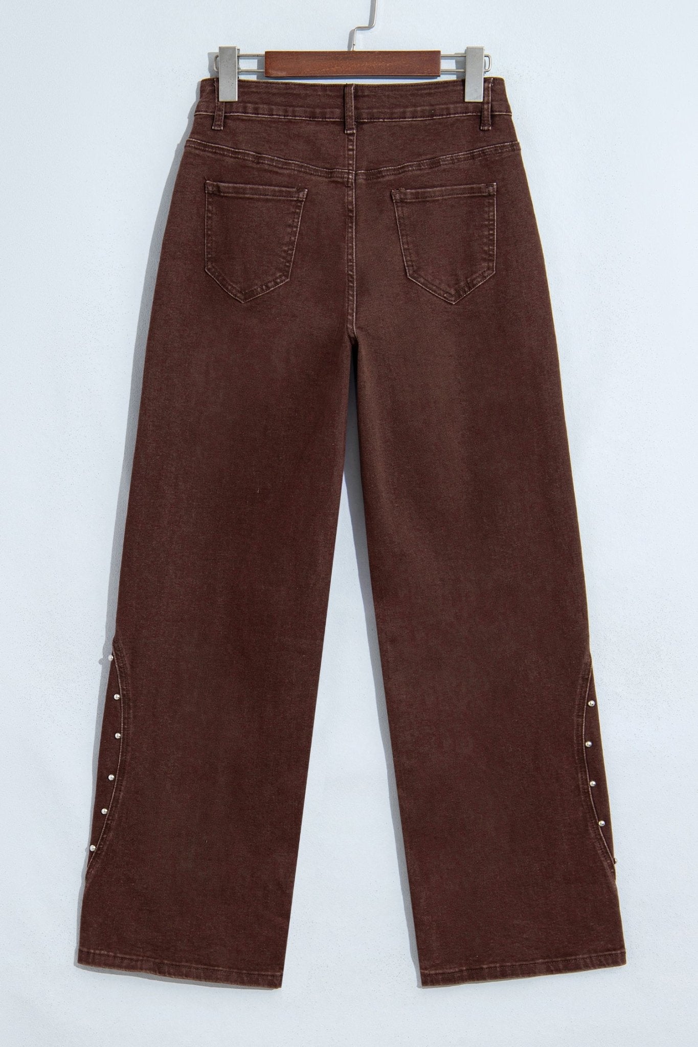 Dark brown wide-leg jeans with silver studs along the outer seams on a wooden hanger
