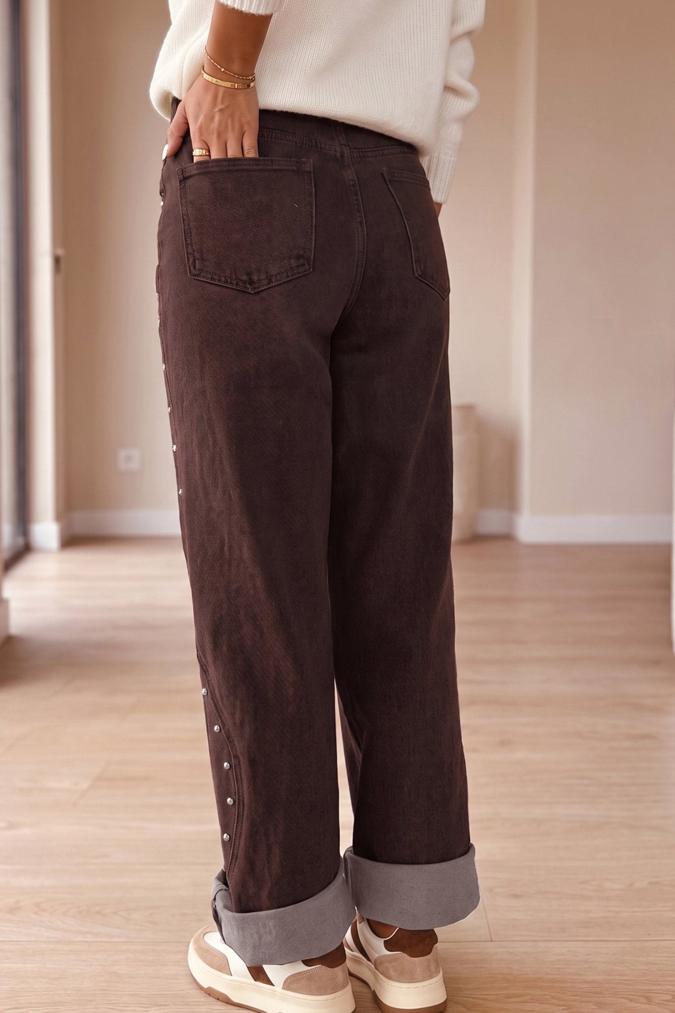 Back view of dark brown wide-leg cuffed jeans with side buttons, cream sweater, and white sneakers.