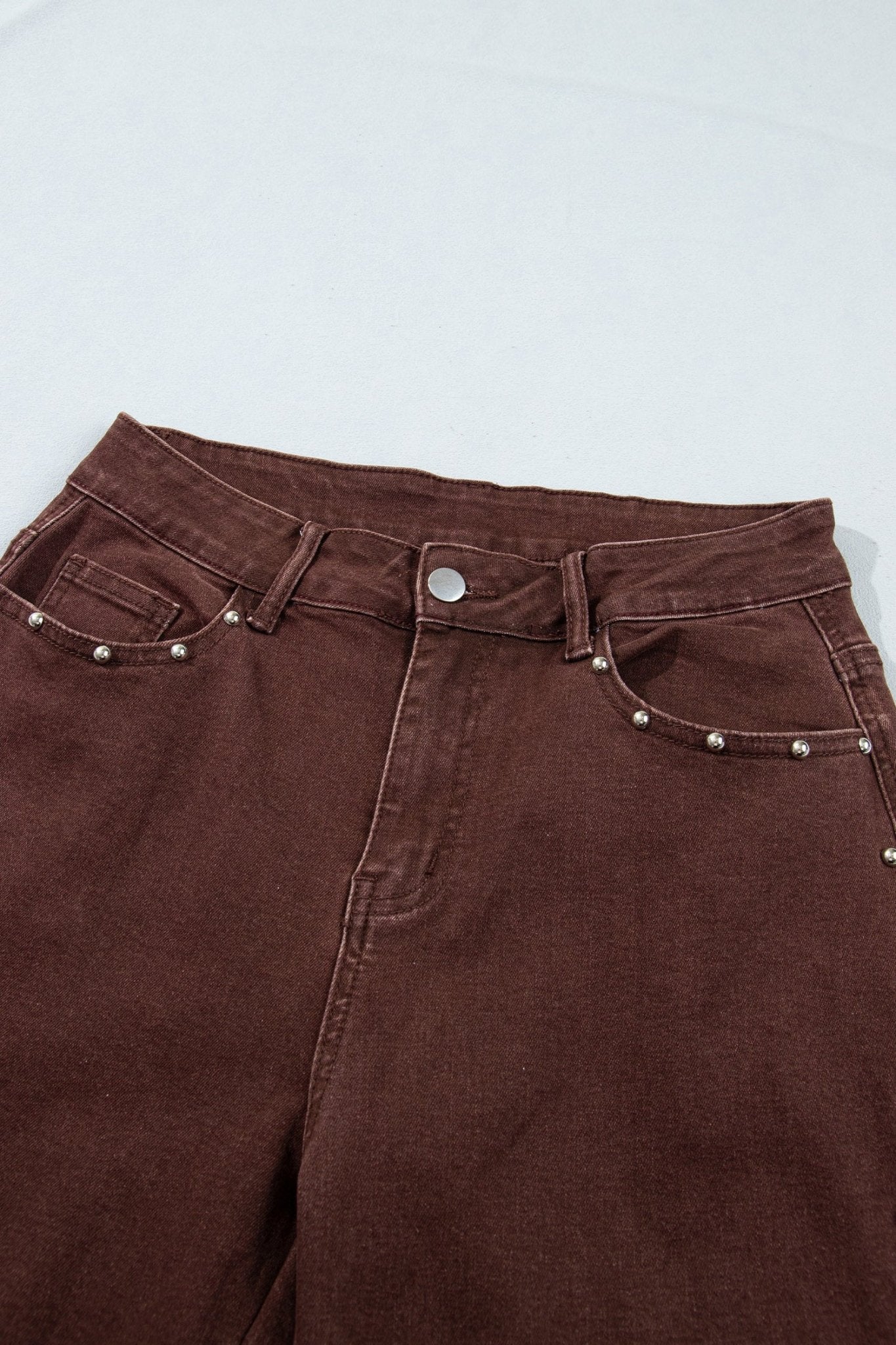 Close-up of dark brown jeans with waistband, pockets, button, and silver rivets