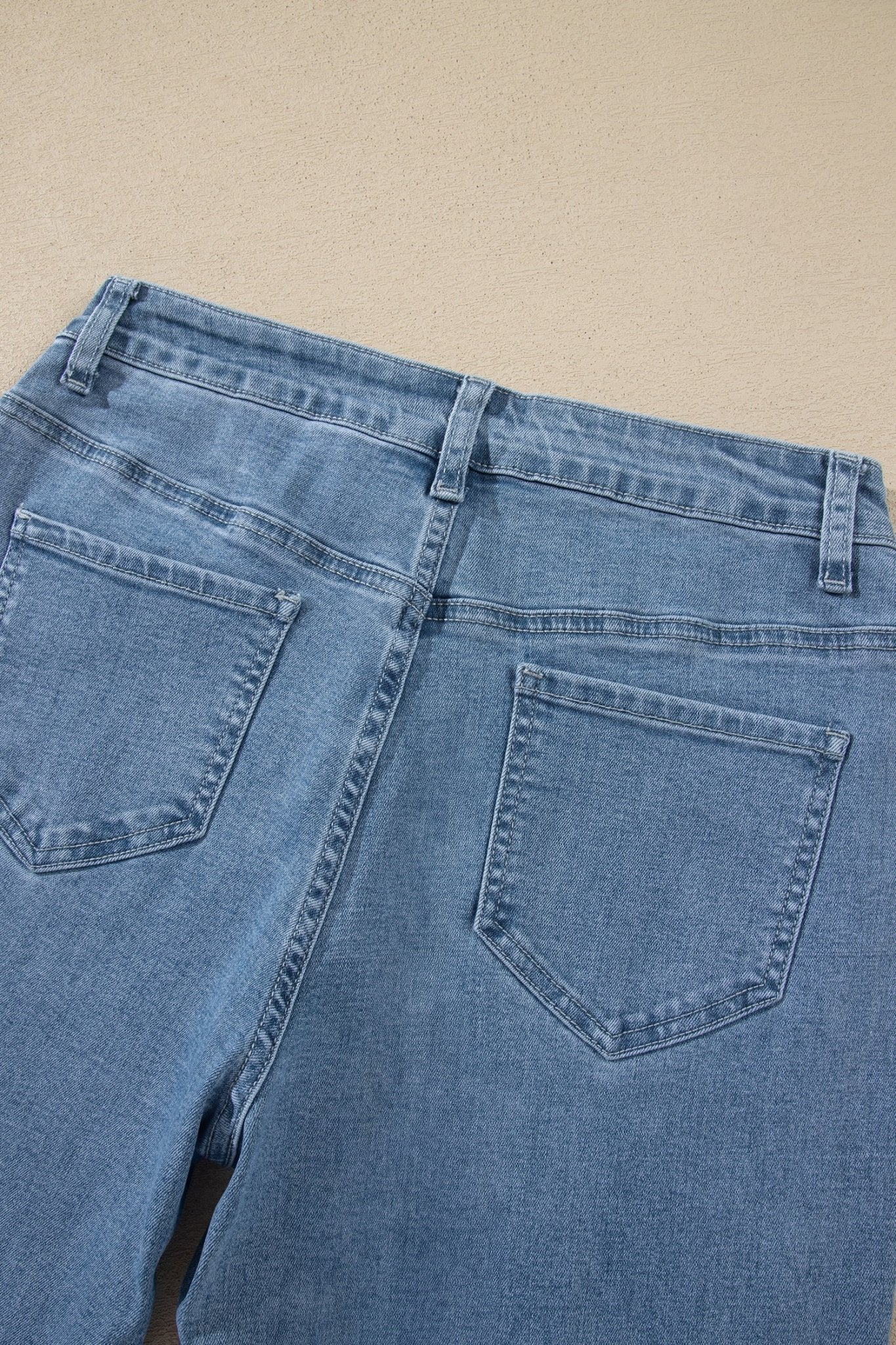 Close-up of the back waistband and pockets of light blue denim jeans laid flat on a beige surface.