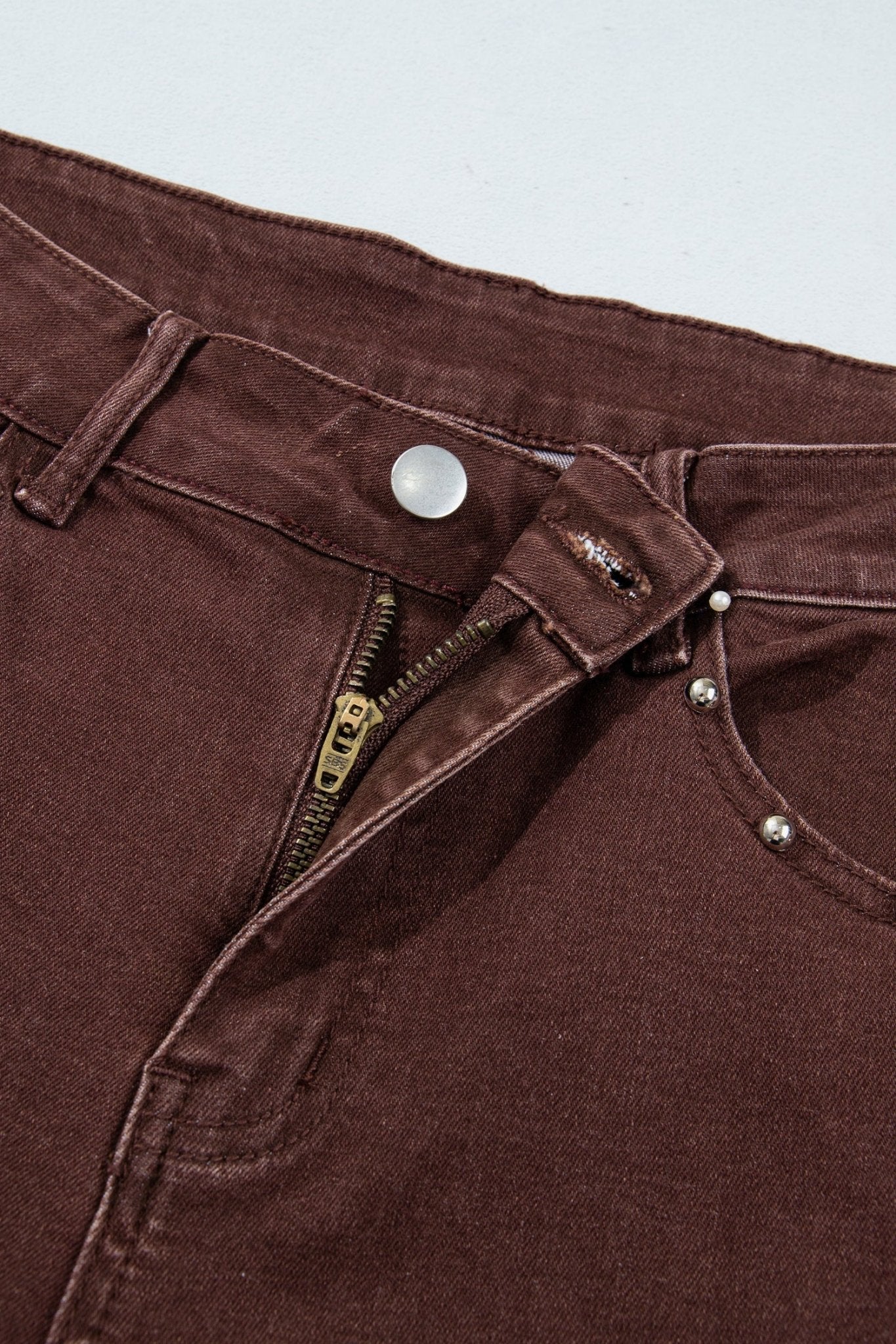 Close-up of dark brown denim pants with waistband, zipper, rivets, and pockets.