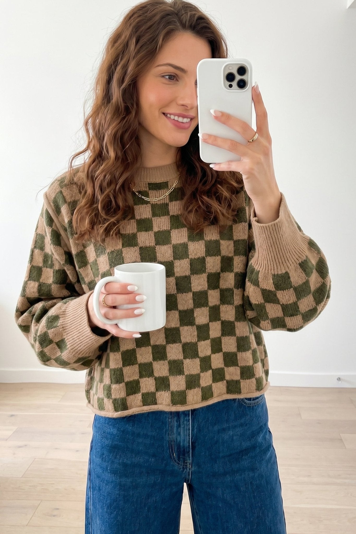 A woman takes a mirror selfie wearing a brown and olive green checkered knit sweater.