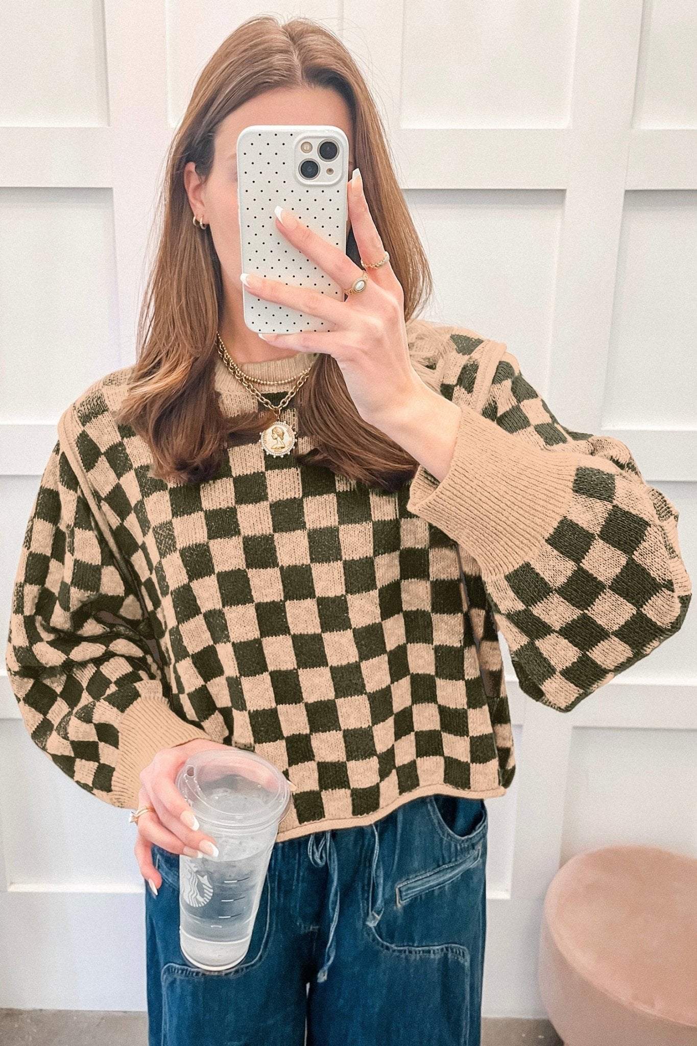 Woman taking mirror selfie in beige and dark green checkered knit sweater with balloon sleeves and blue jeans.