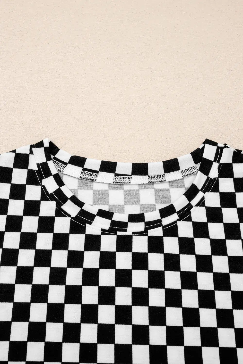 A close-up of a black and white checkerboard patterned top, showing the crew neck and inner collar label, laid flat against a light beige background.