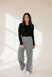 Woman wearing a black top and checkered pants standing against a beige wall.