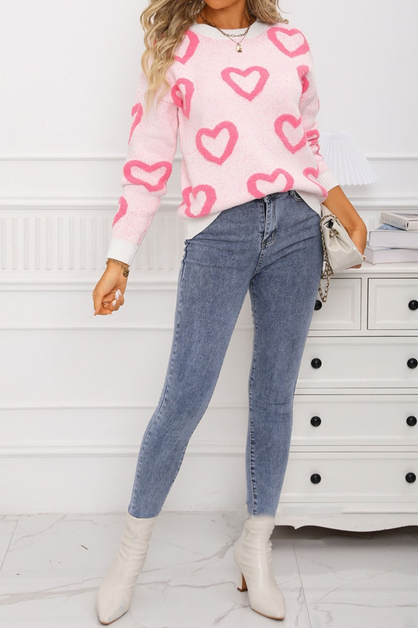 Person wearing a pink sweater with heart patterns, blue jeans, and white boots in a room.