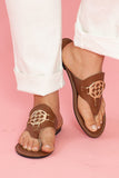 Brown sandals with a gold logo worn by a person on a pink background