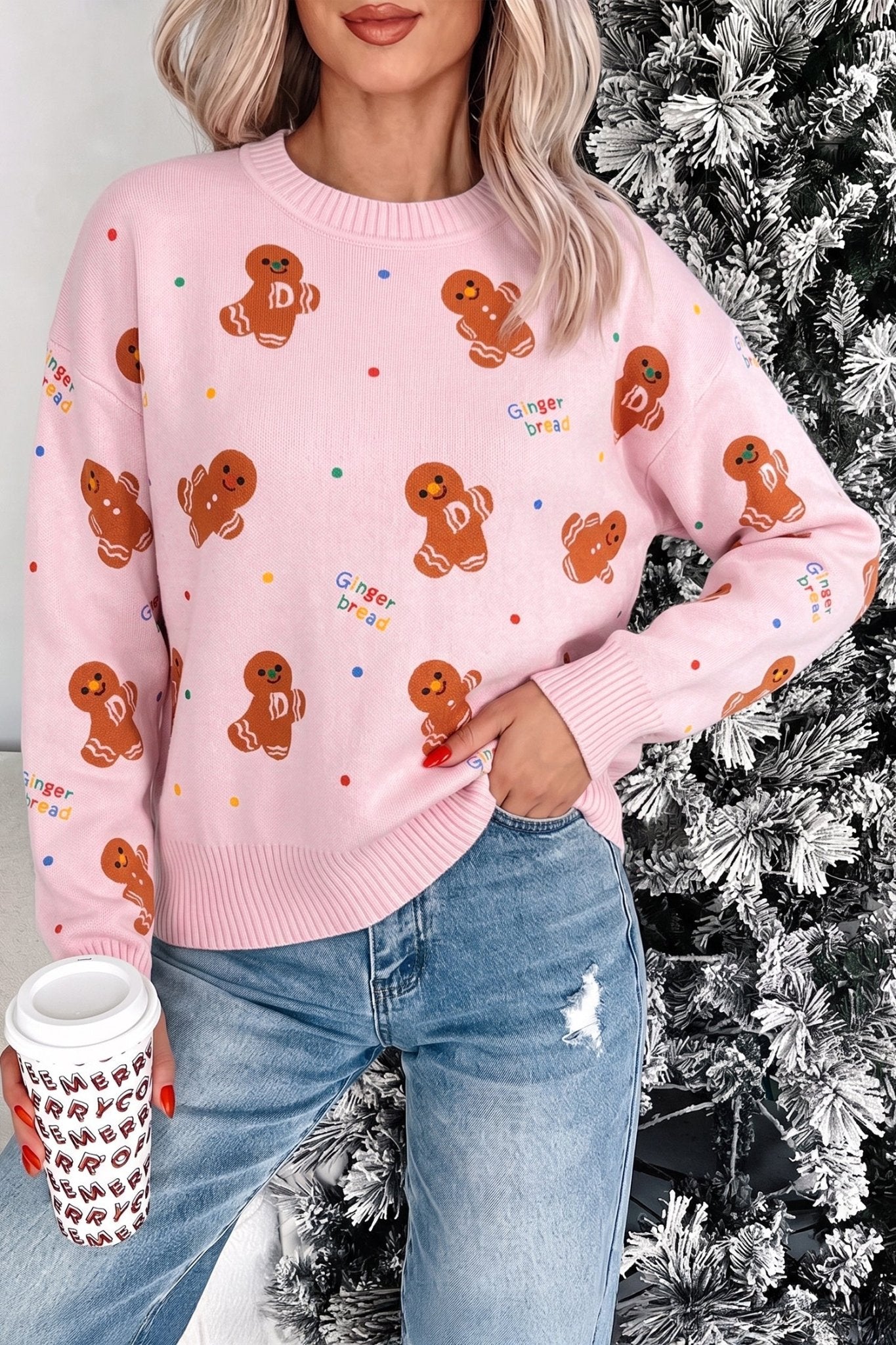 Woman in pink gingerbread-print sweater with blue jeans holding coffee by Christmas tree.
