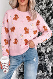 Woman in pink gingerbread-print sweater with blue jeans holding coffee by Christmas tree.