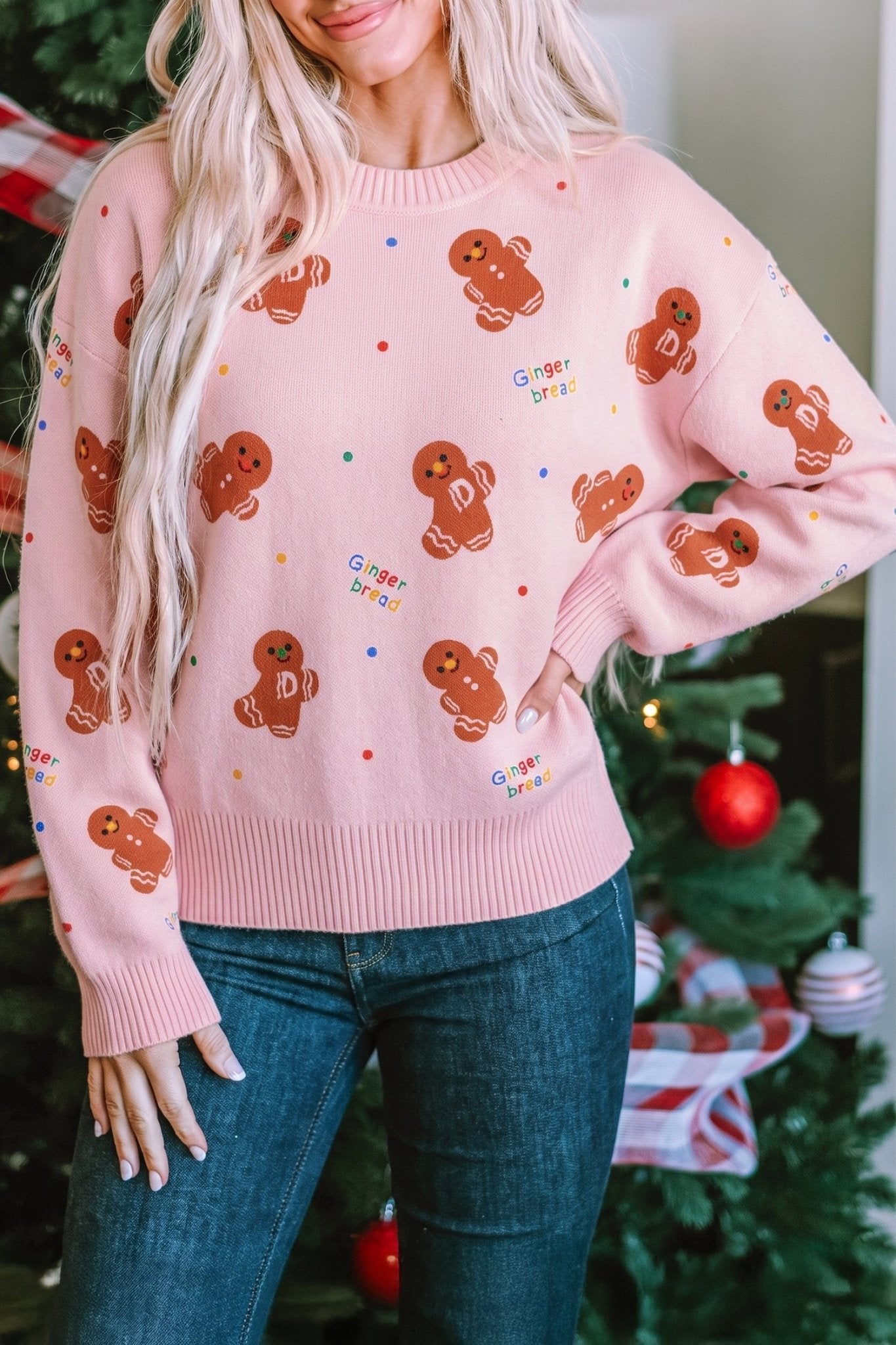 Woman in light pink gingerbread-pattern sweater with dark blue jeans by Christmas tree.
