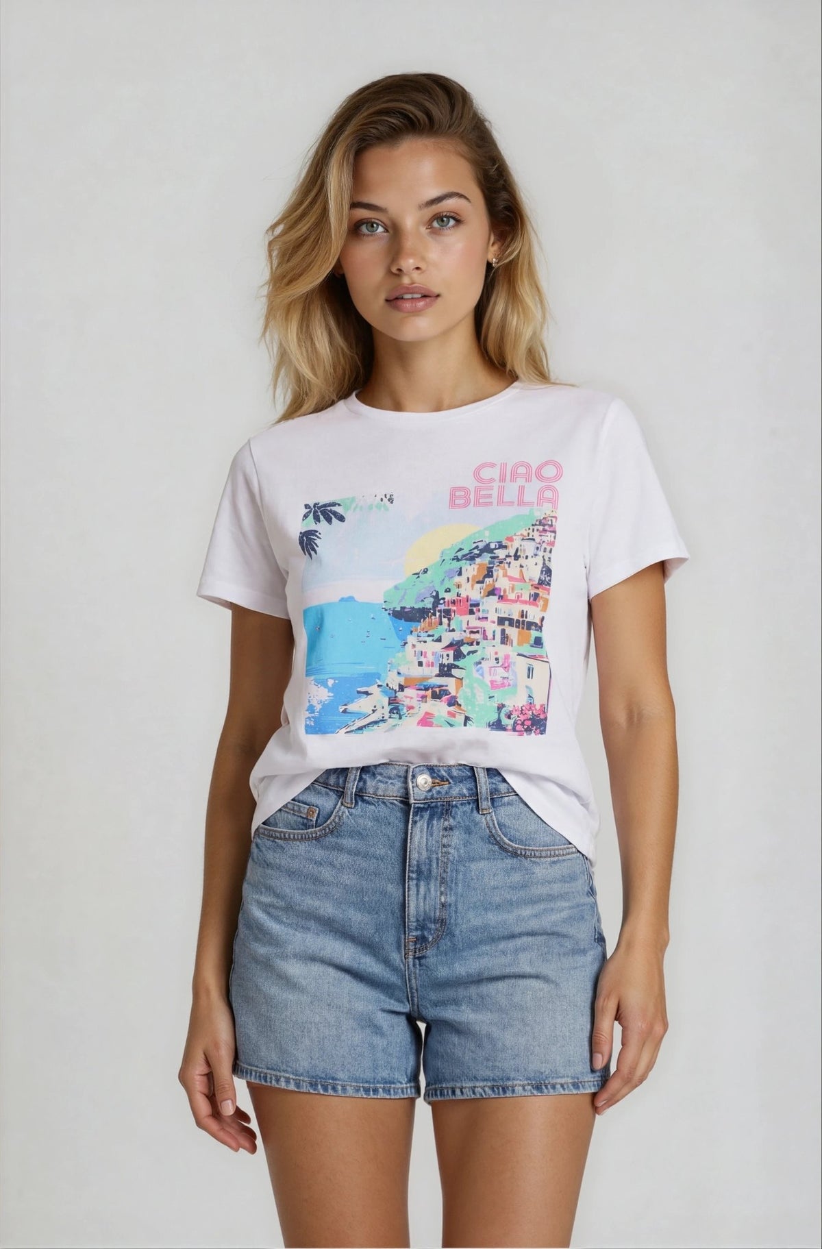 Woman wearing a white t-shirt with colorful graphic and 'Ciao Bella' text, paired with denim shorts on a plain background.