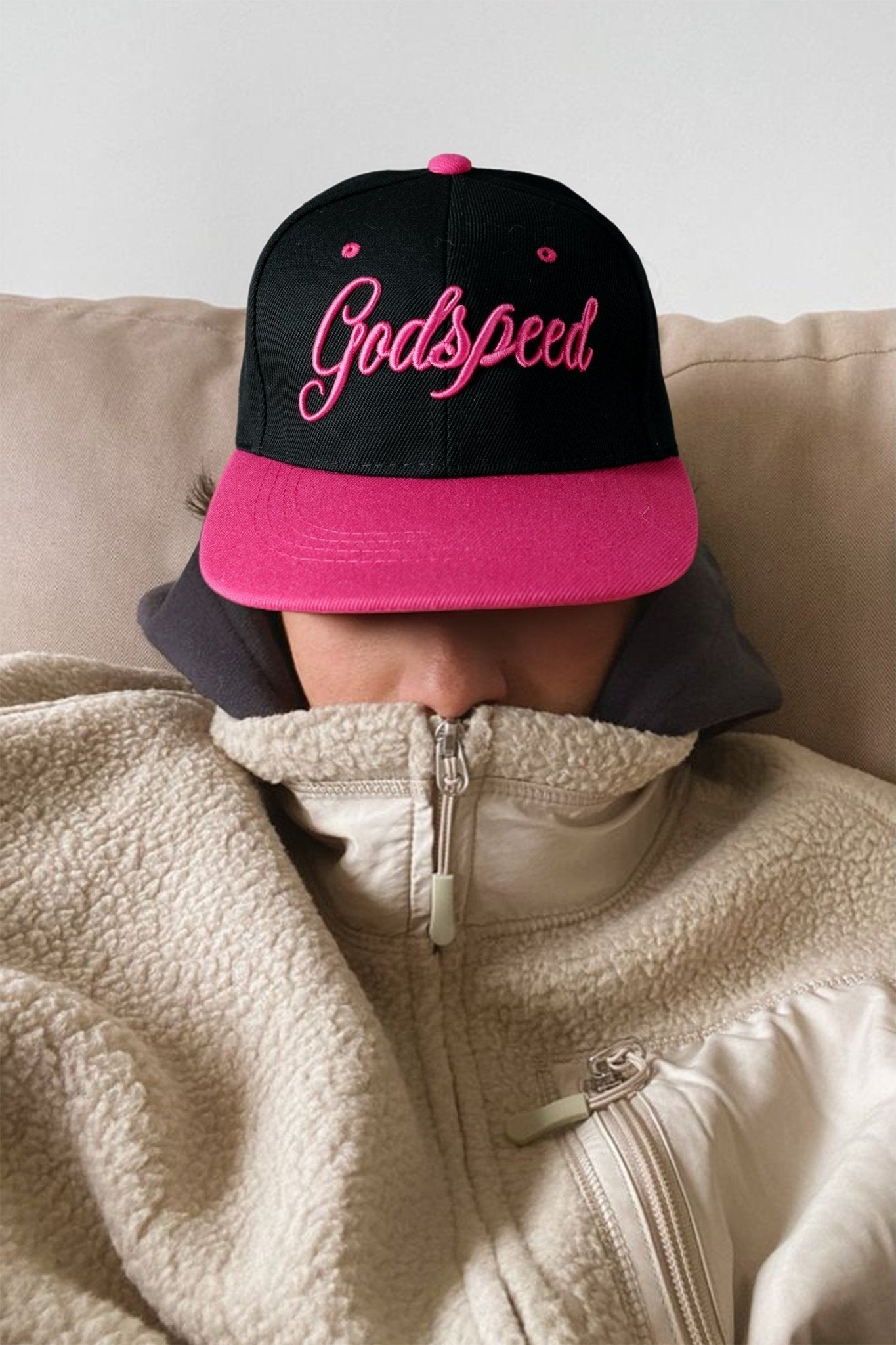 Person wearing a black and pink 'Godspeed' cap and beige fleece jacket on a couch.
