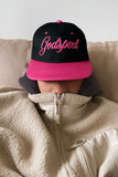 Person wearing a black and pink 'Godspeed' cap and beige fleece jacket on a couch.